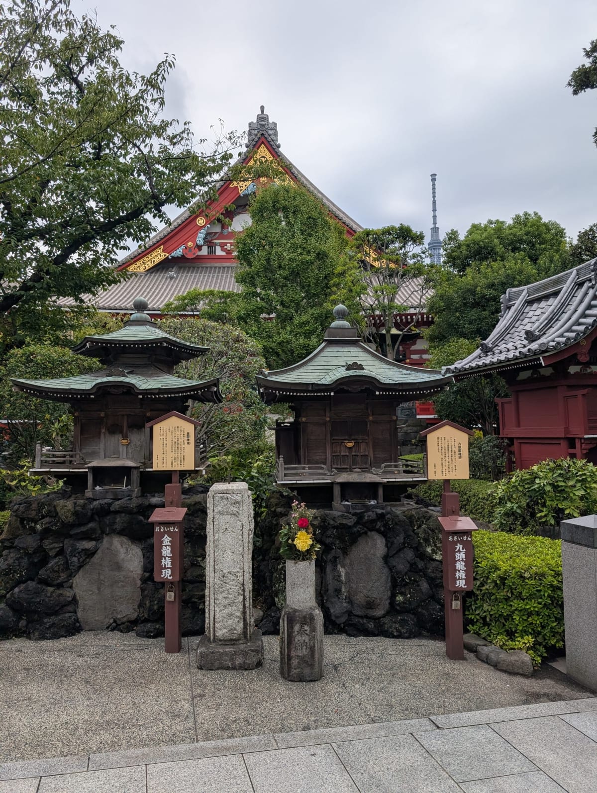 Two small copper-roofed shrine buildings, Kinryu-gongen and Kuzuryu-gongen, on a bed of volcanic rock at Senso-ji, with Tokyo Skytree visible through the trees behind them