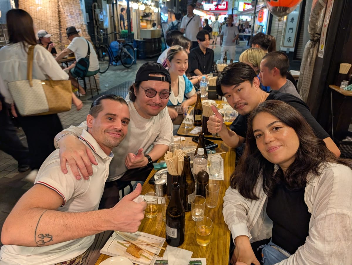 A group photo around a crowded izakaya table in Ueno's market alleys at night, Pierre and new friends smiling and giving thumbs up, bottles and chopsticks covering the table