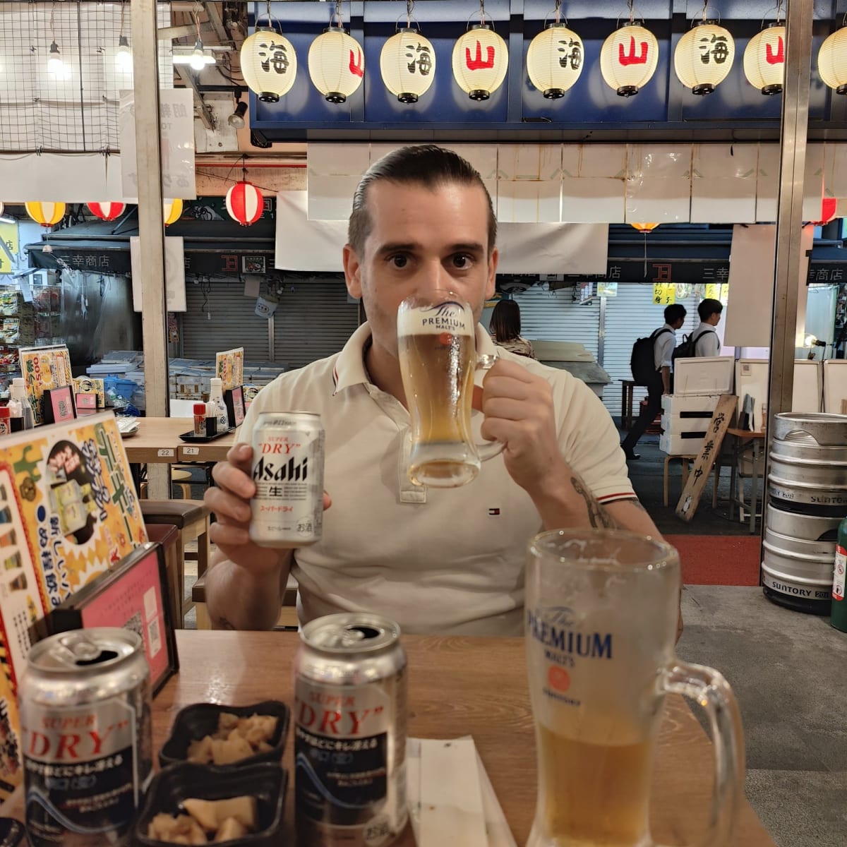 Pierre holding a can and mug of Asahi Super Dry beer at an outdoor izakaya set up among Ameyoko's shuttered fish stalls, lanterns reading umi and yama overhead