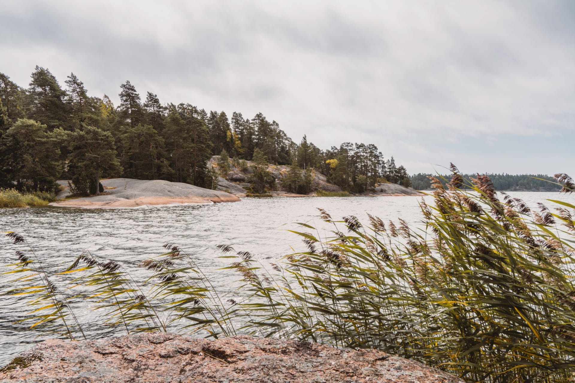Low granite ruins of a 19th-century Russian fortress scattered across a green shoreline meadow beside calm Baltic water on Åland, with birch trees framing the broken walls.