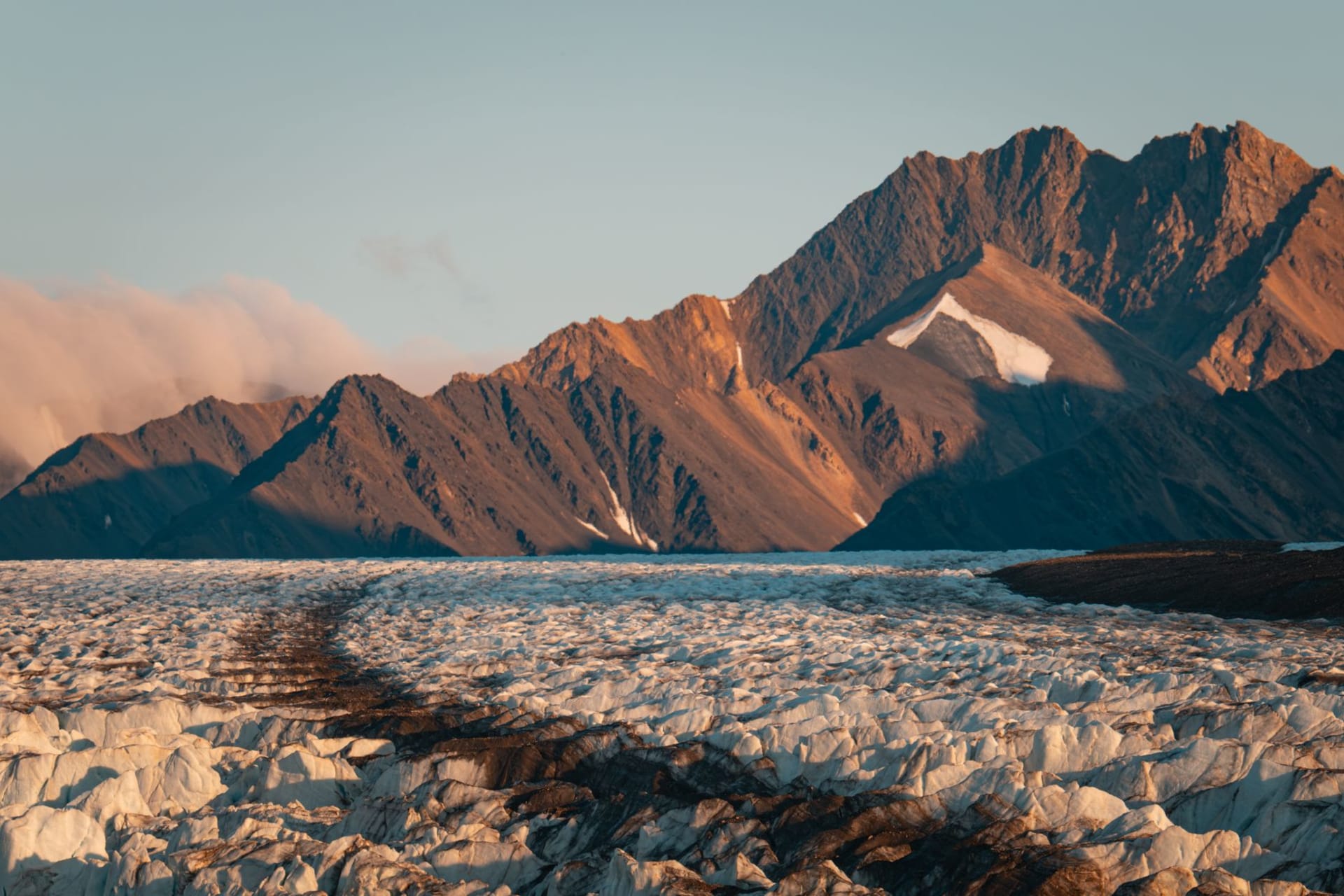 Dramatic glacial and mountain landscape bathed in golden hour light, with jagged peaks rising above a vast valley of ice and snow