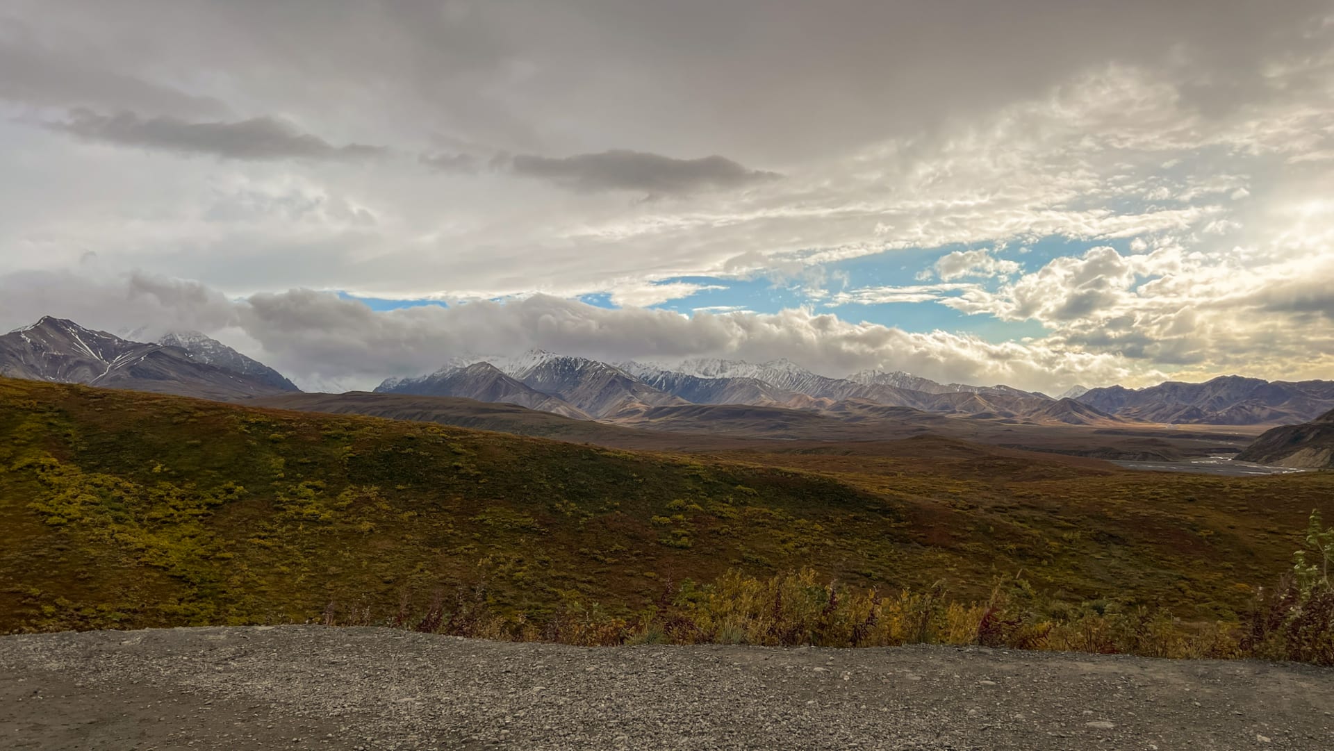 Denali's massive snow-covered summit rising above clouds and a vast tundra valley under summer light