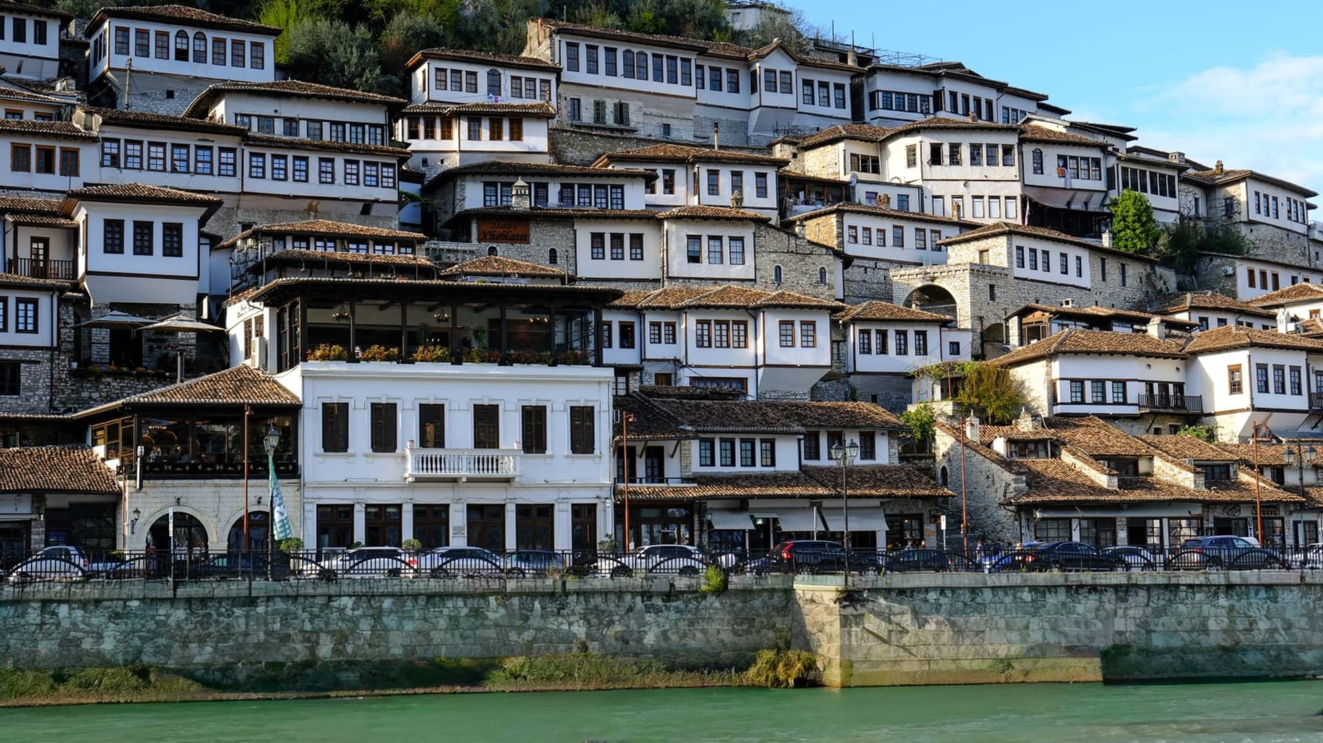 White Ottoman-era houses stacked in dense tiers on a steep hillside above the green Osum river, their rows of tall arched windows catching afternoon light in Berat, Albania.
