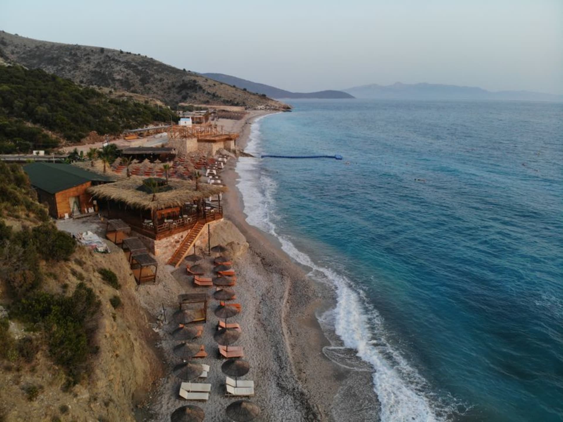 La playa de guijarros de Dhërmi curvándose junto al mar Jónico turquesa bajo montañas verdes empinadas, en la Riviera de Albania.
