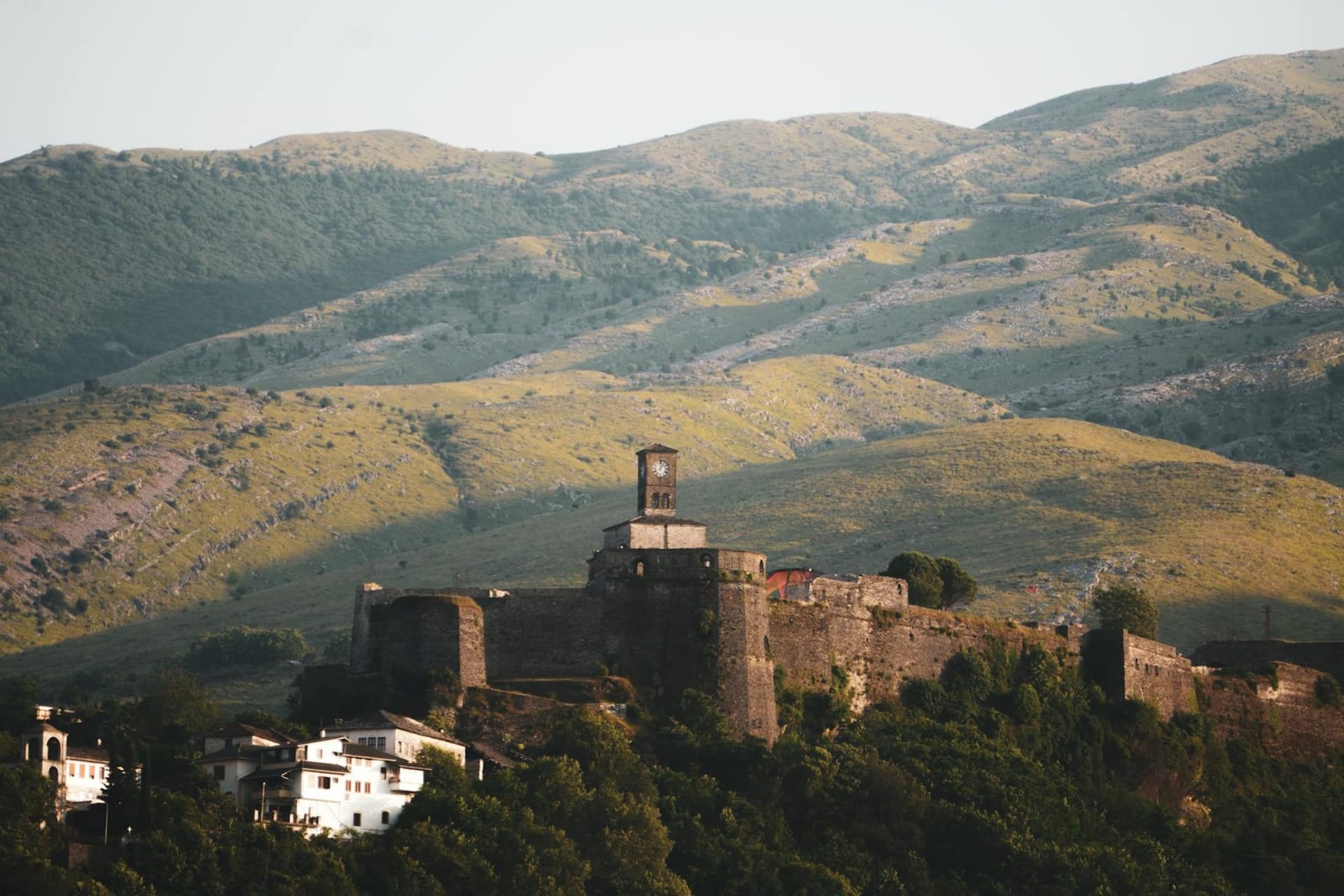 Stone tower houses with slate rooftops rising along a steep hillside in Gjirokaster, Albania, with the massive Ottoman castle looming above against a grey mountain sky.