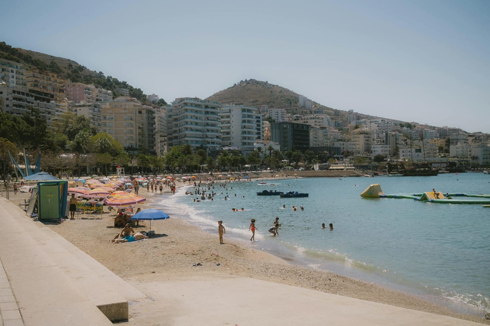 Saranda's curved seafront promenade hugging the turquoise Ionian bay, white and terracotta buildings stacked up the hillside behind it, with the green outline of Corfu visible across the water in the late afternoon haze.
