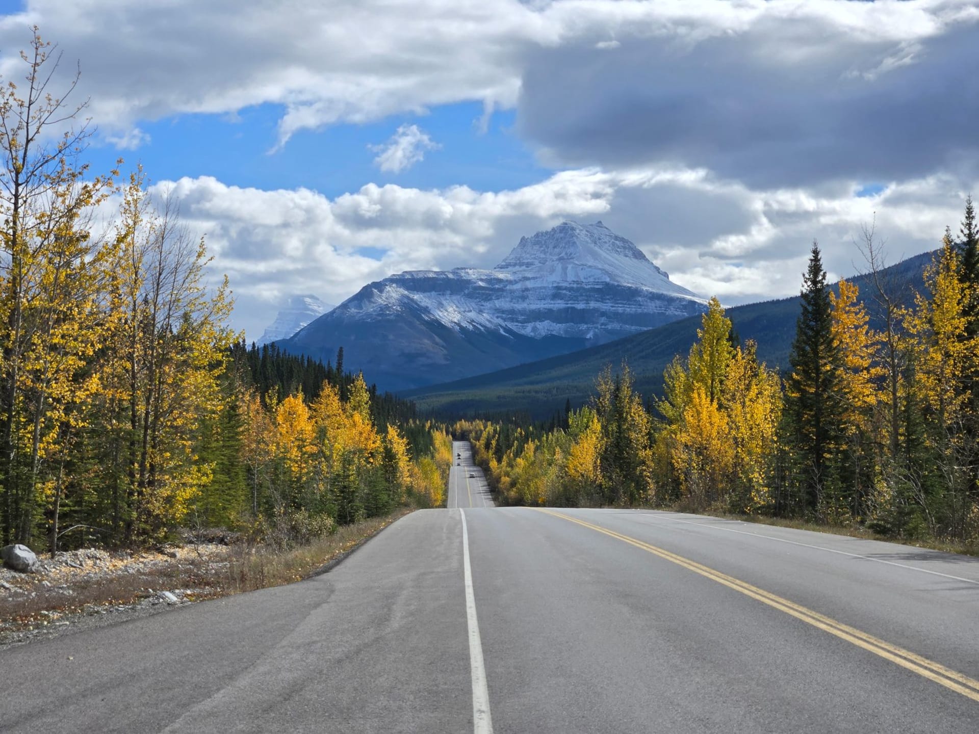The Icefields Parkway stretching into the distance between towering rock walls and a turquoise glacial river, a single car visible far ahead