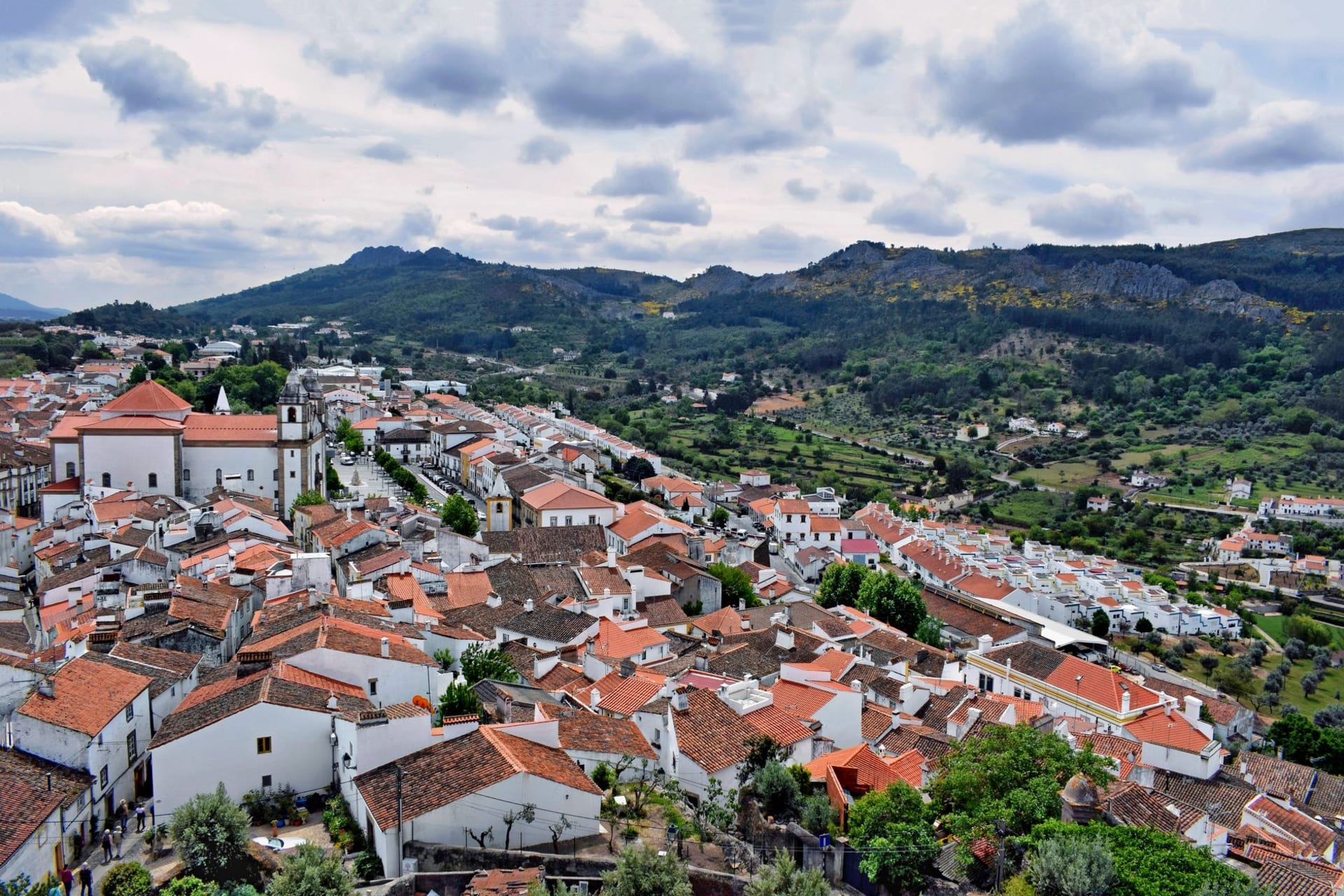 A tiny whitewashed hilltop village ringed by medieval stone walls and crowned by a castle, perched on a high granite crag above a vast green plain in Marvão, Alentejo, Portugal.