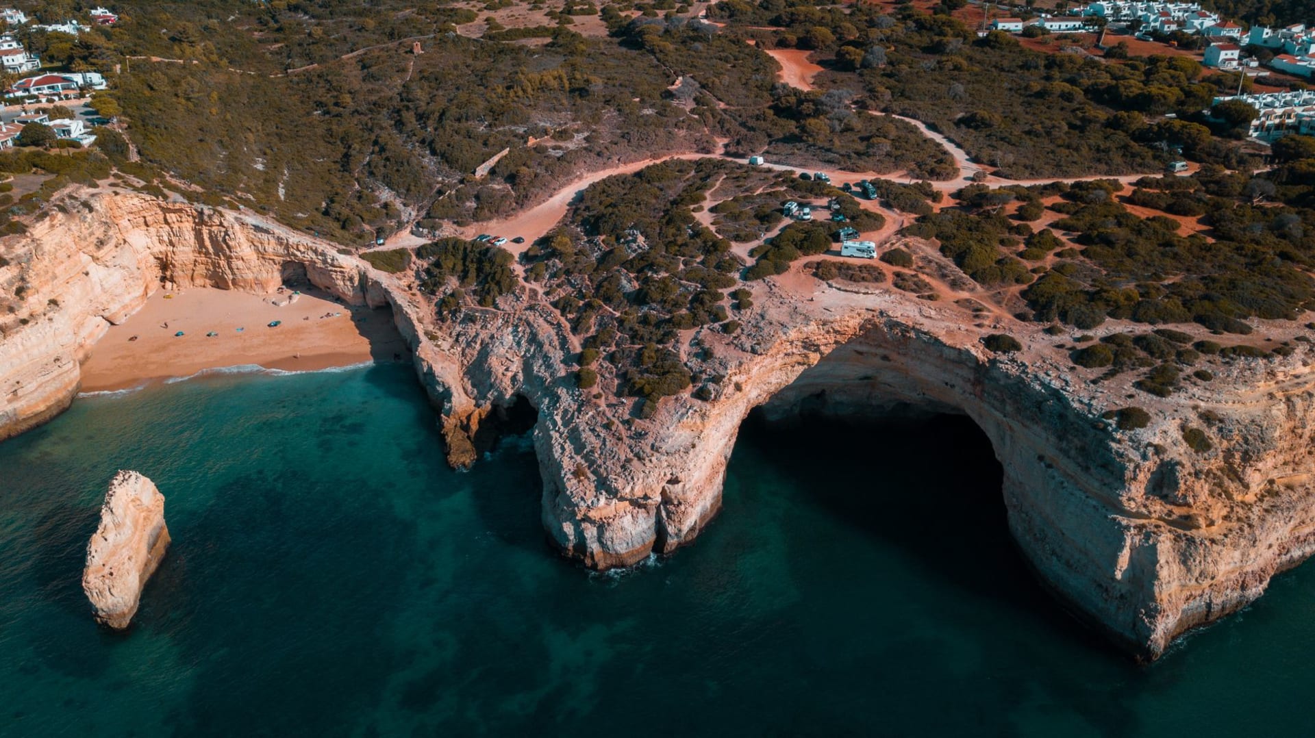 Aerial view of twin sea arches carved into golden sandstone cliffs at the Algarve coast, with turquoise water below and a sandy cove beach alongside