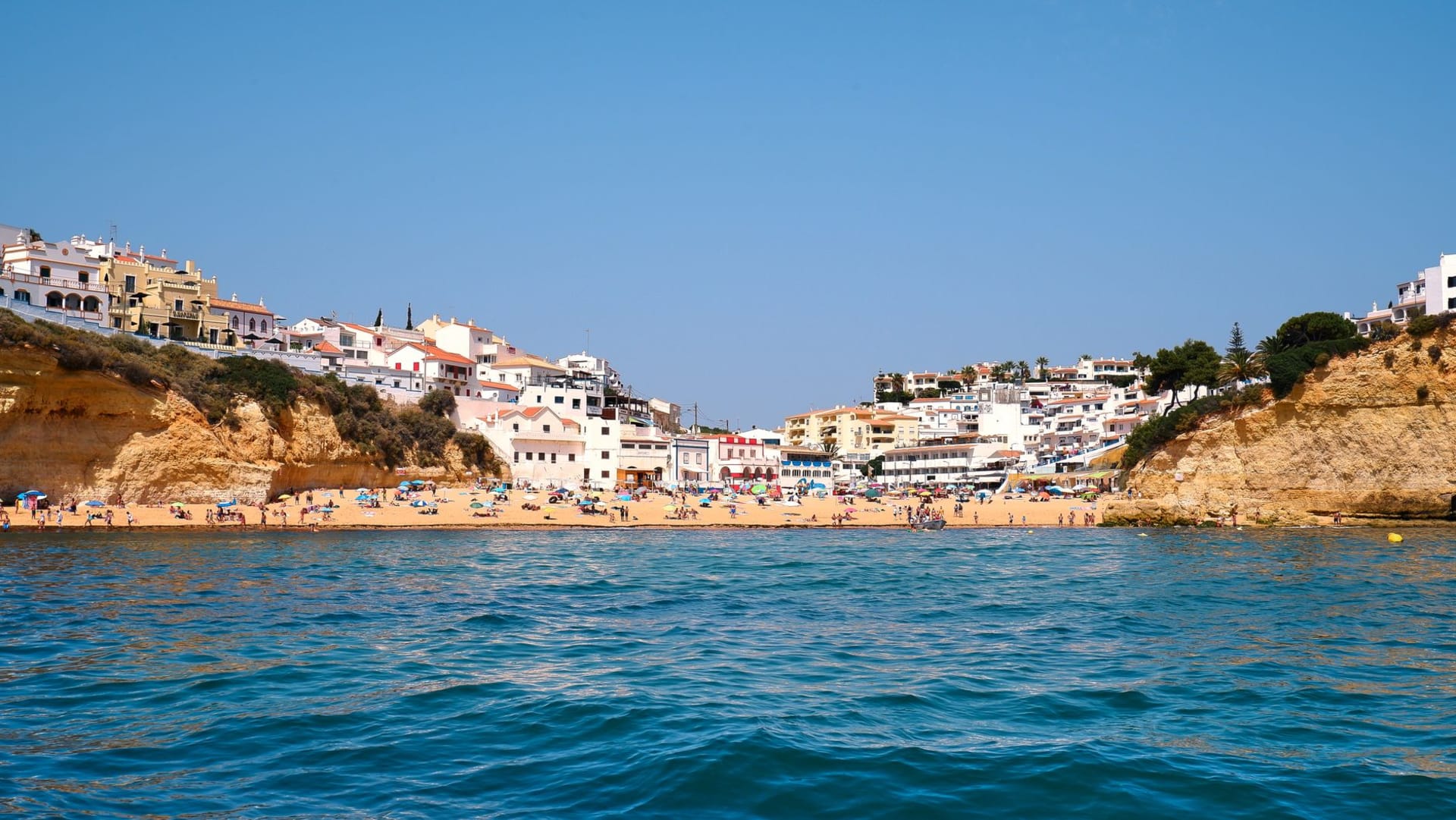 Carvoeiro village beach seen from above — a small sandy cove enclosed by golden sandstone cliffs, colorful fishing boats and whitewashed houses framing the water
