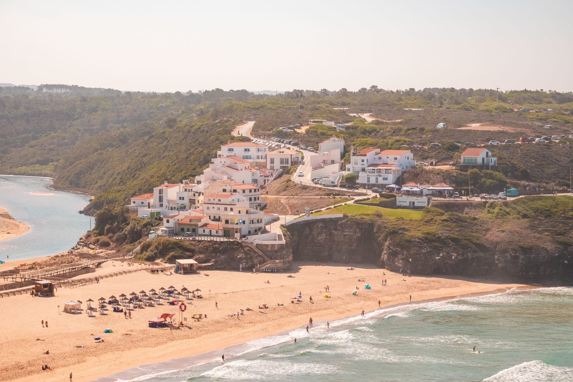 Praia de Odeceixe where the Seixe river meets the Atlantic, seen from the clifftop — a wide sandy beach enclosed by golden cliffs with the river cutting through the sand