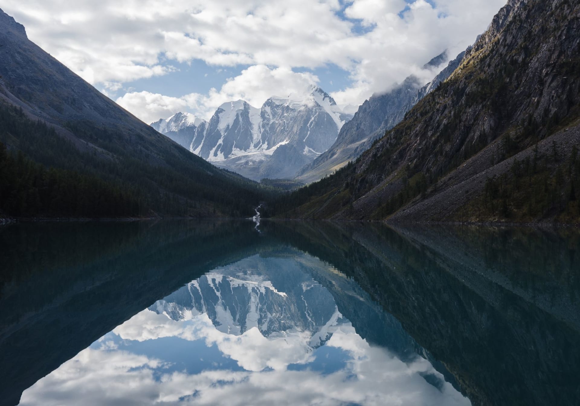 A perfectly still glacial lake in the Altai Mountains reflecting snow-capped peaks and dramatic clouds between steep forested valley walls