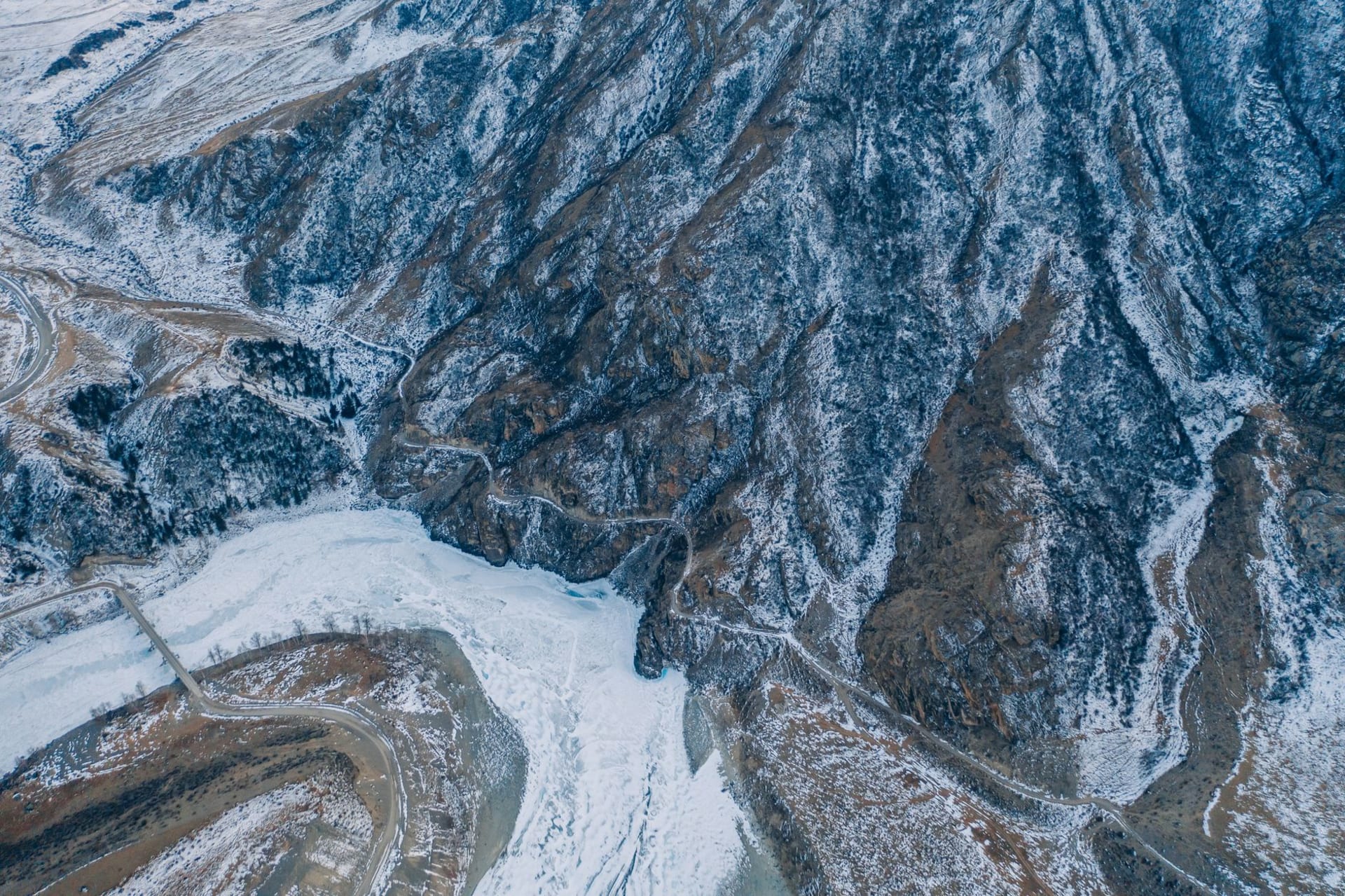 The Chuya Highway cutting through a wide mountain valley with turquoise river below, steep canyon walls on both sides, and snow-capped peaks at the far end