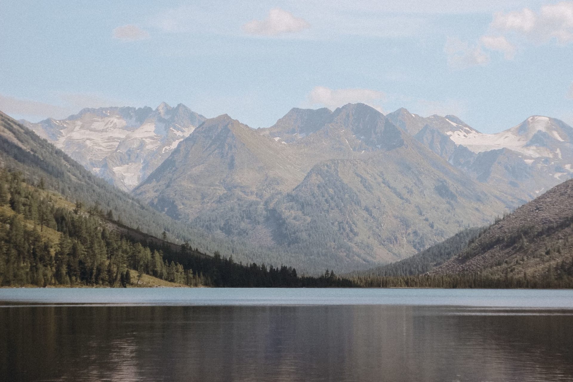 Deep blue Lake Teletskoye stretching between steep forested mountain ridges under a clearing sky, with a small wooden boat near the dark shoreline in the Russian Altai.