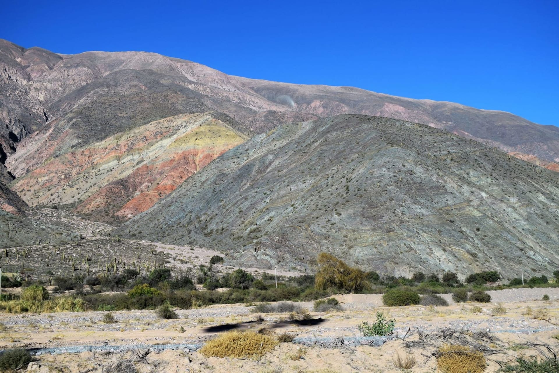 A sweeping view of the Quebrada de Humahuaca gorge at midday, with striated Andean mountains glowing in layers of ochre, burgundy, violet, and pale green beneath a deep blue sky.