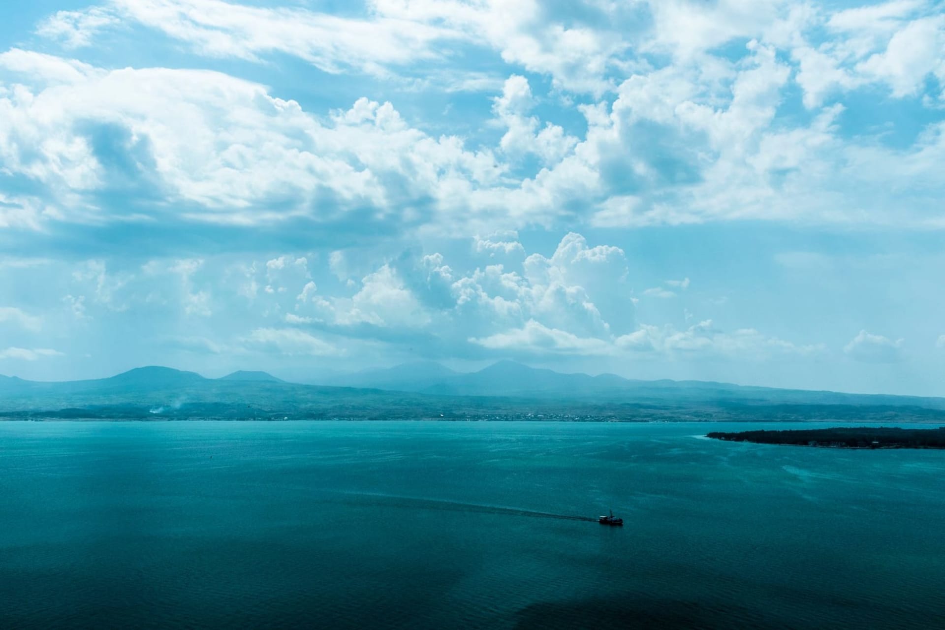 A vast turquoise lake stretching to the horizon under an enormous open sky, with the stone towers of Sevanavank monastery rising from a rocky peninsula in the foreground