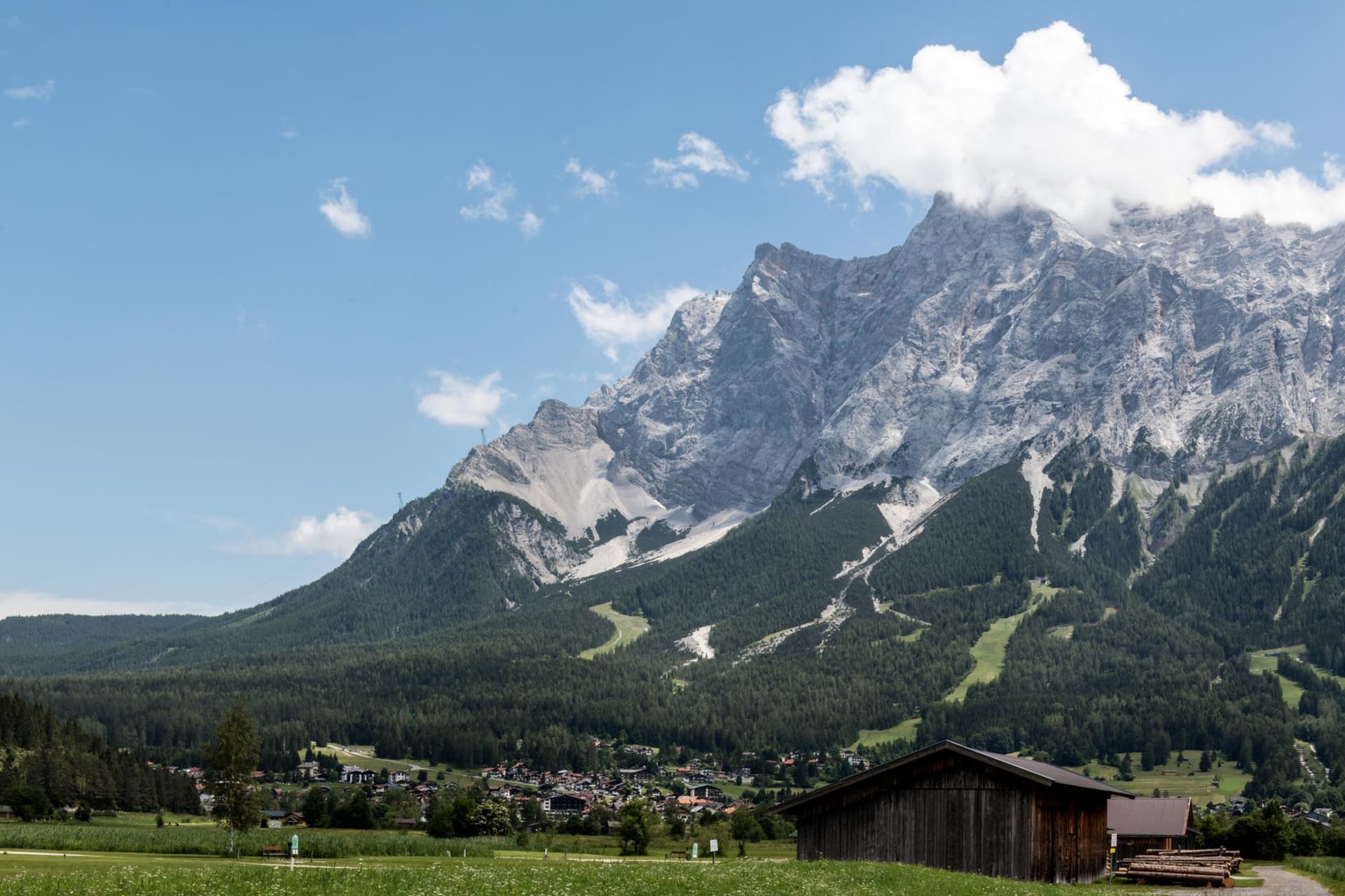 A traditional wooden barn in a green alpine meadow with the jagged grey limestone massif of the Zugspitze rising above the forested slopes and a small Tyrolean village below, under a blue summer sky