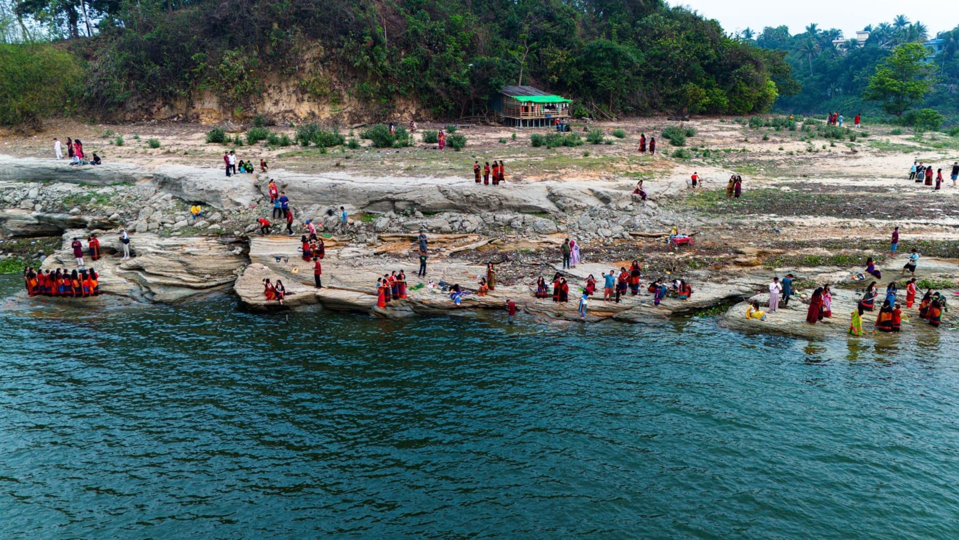 Wooden stilt houses of a Chakma village rising above the glassy Kaptai reservoir at dawn, surrounded by forested hills and a wooden boat docked at a bamboo jetty.