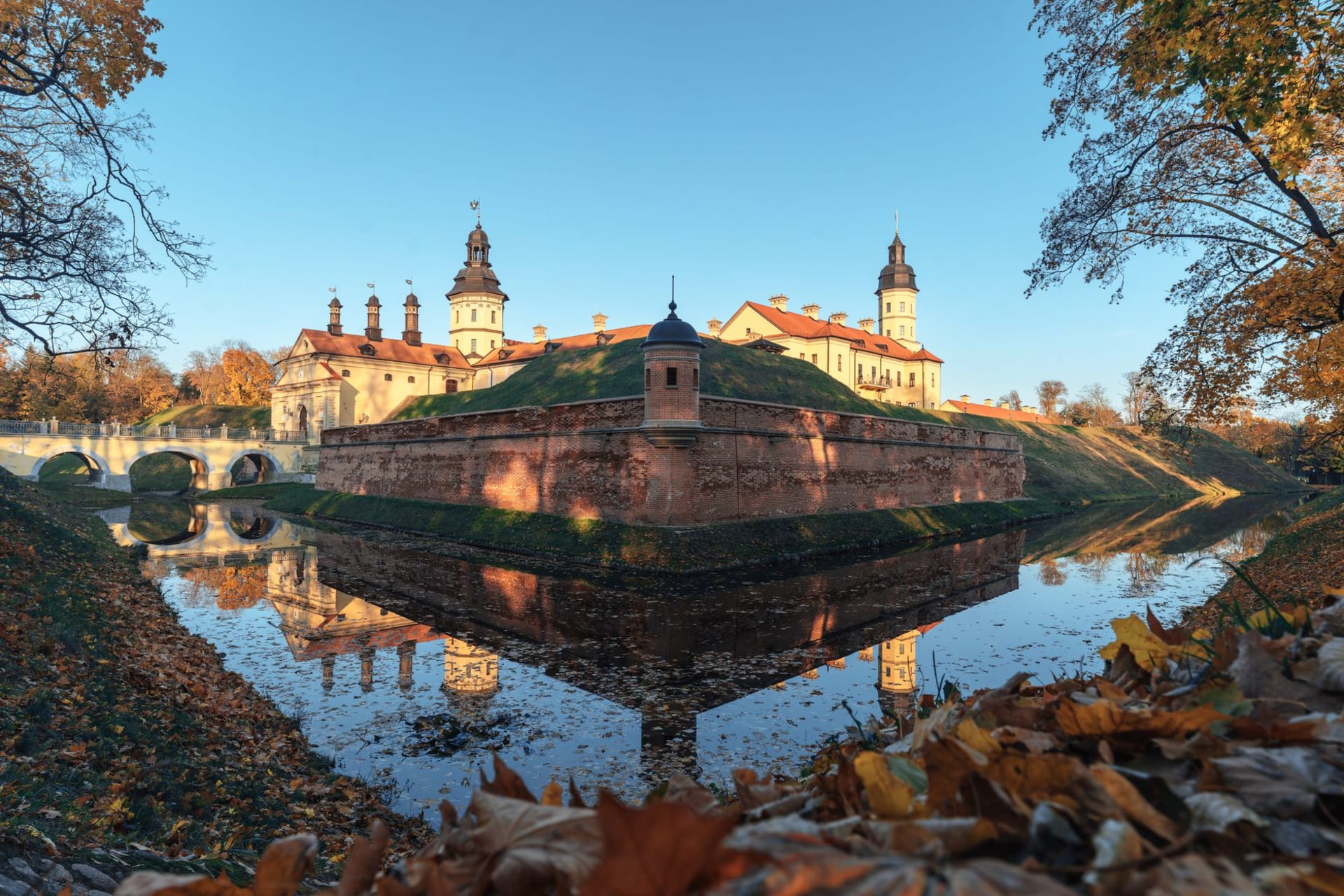 The ornate Baroque façade of Nesvizh Palace reflected in a formal garden pond, autumn parkland surrounding it in shades of gold and russet