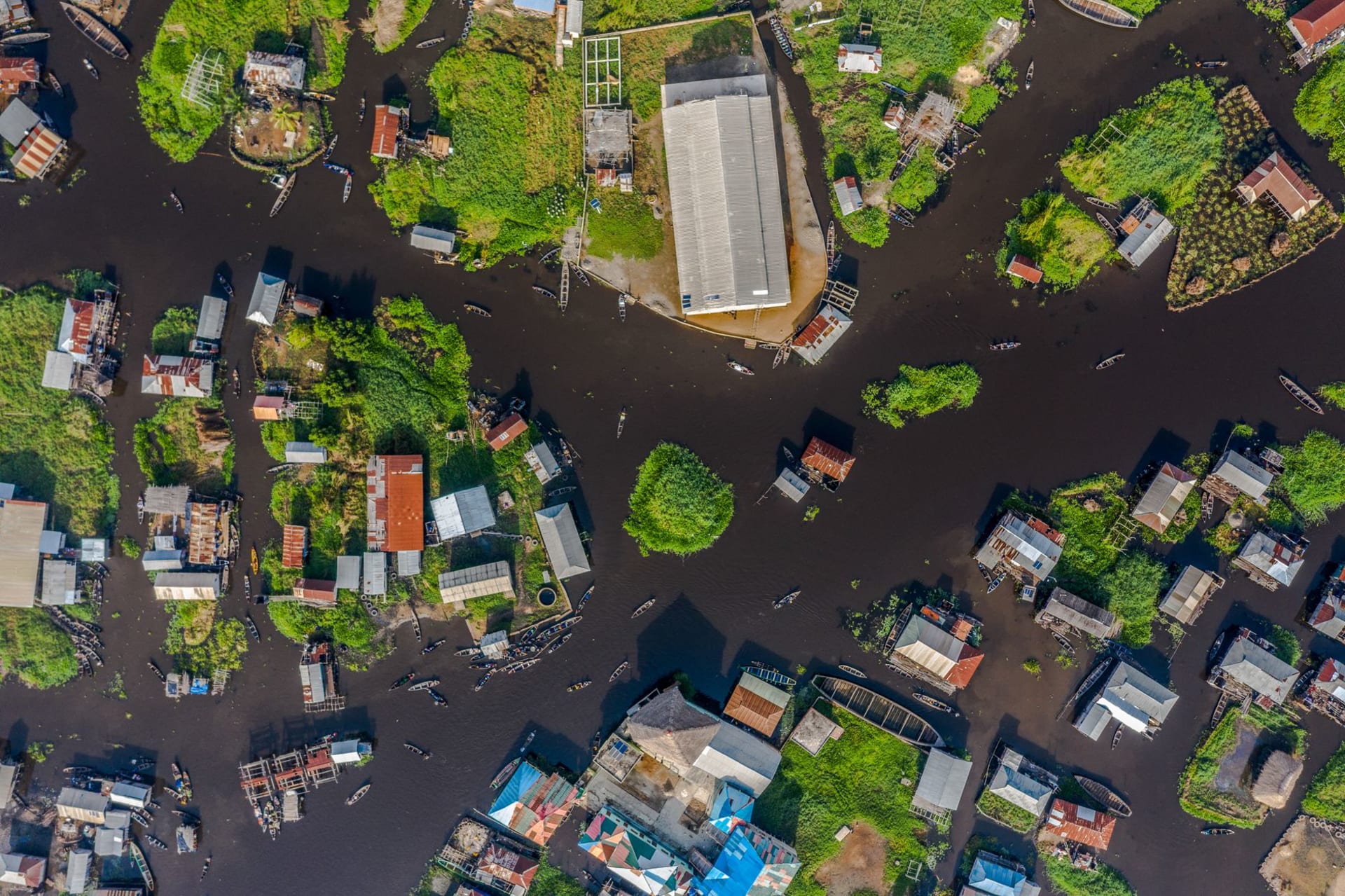 Aerial drone view of Ganvié stilt village on Lake Nokoué, with wooden houses rising from brown water and dugout canoes threading between them