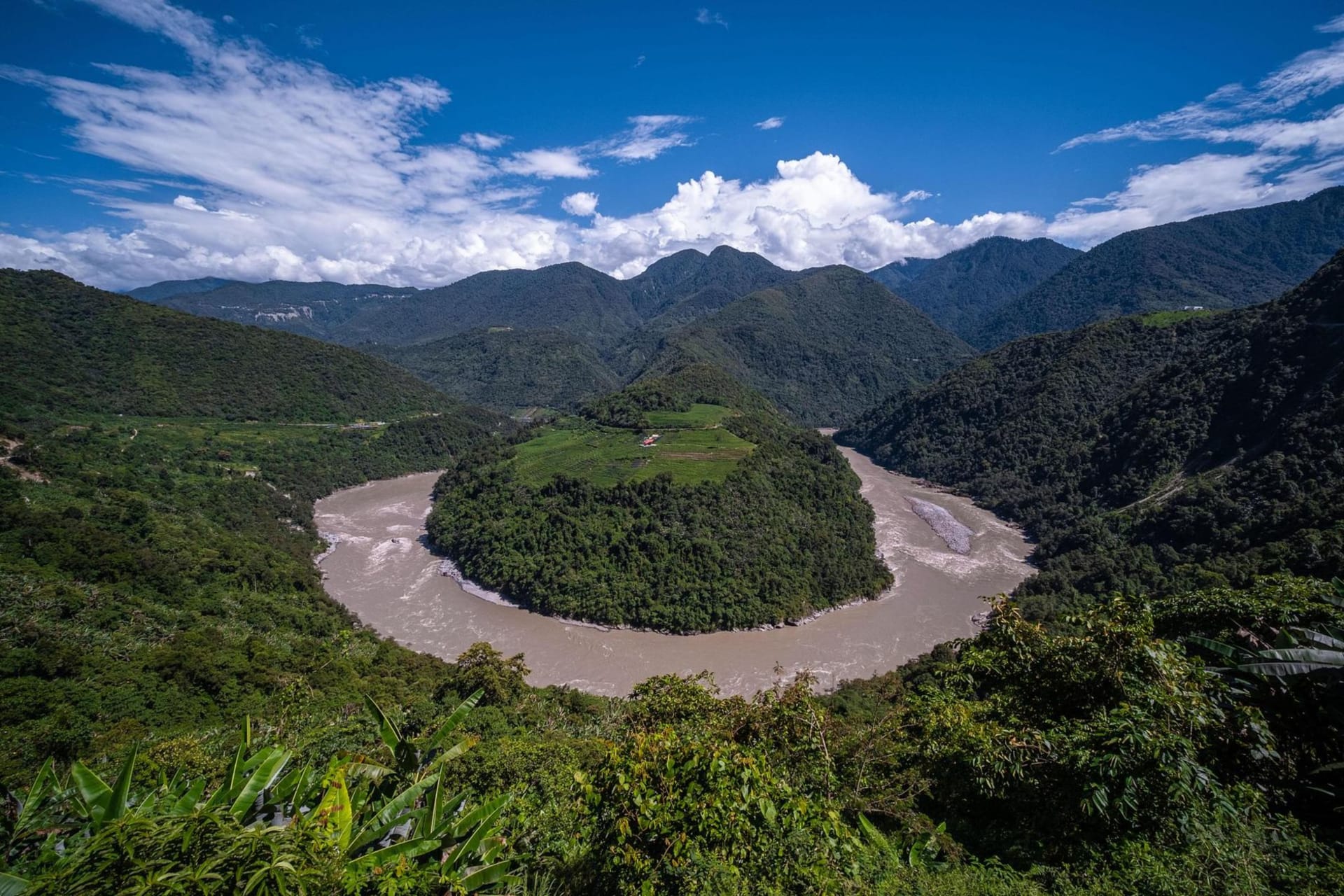 A narrow mountain road carved into a steep cloud-forested slope in the Yungas, mist rising from the valley far below, waterfalls threading the rock face