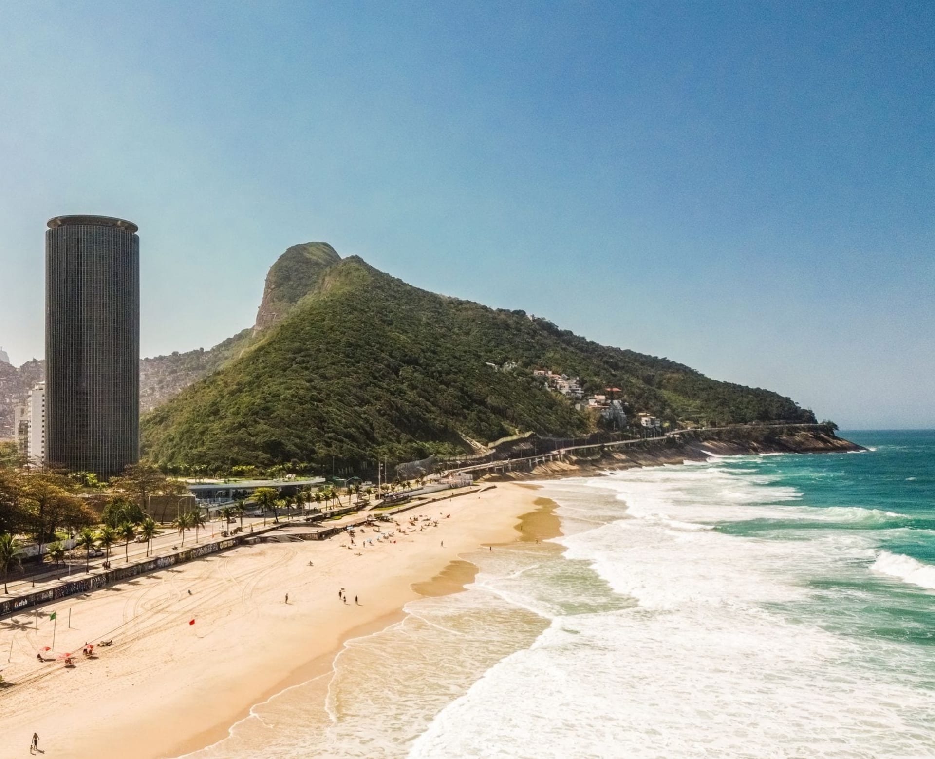 Golden afternoon light falling across the crowded sand of Ipanema beach, with the twin peaks of Morro Dois Irmãos rising sharply above the waterline in the background