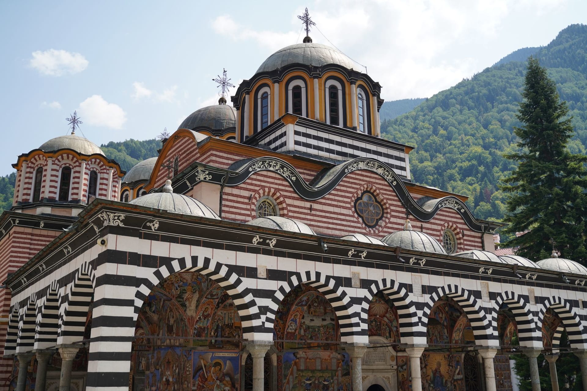 Arcadas pintadas y torre de iglesia abovedada del Monasterio de Rila rodeado de montañas cubiertas de pinos