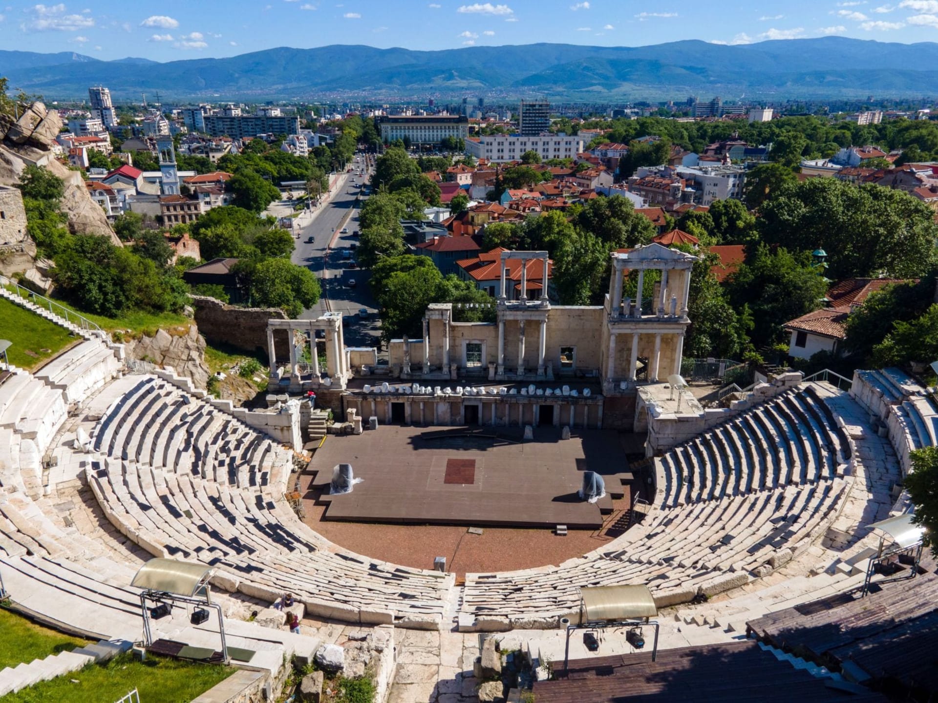 Colorful 19th-century Revival-era houses with ornate painted facades and wooden bay windows cascading down a hillside in Plovdiv's Old Town, with terracotta rooftops and the Rhodope Mountains visible in the distance