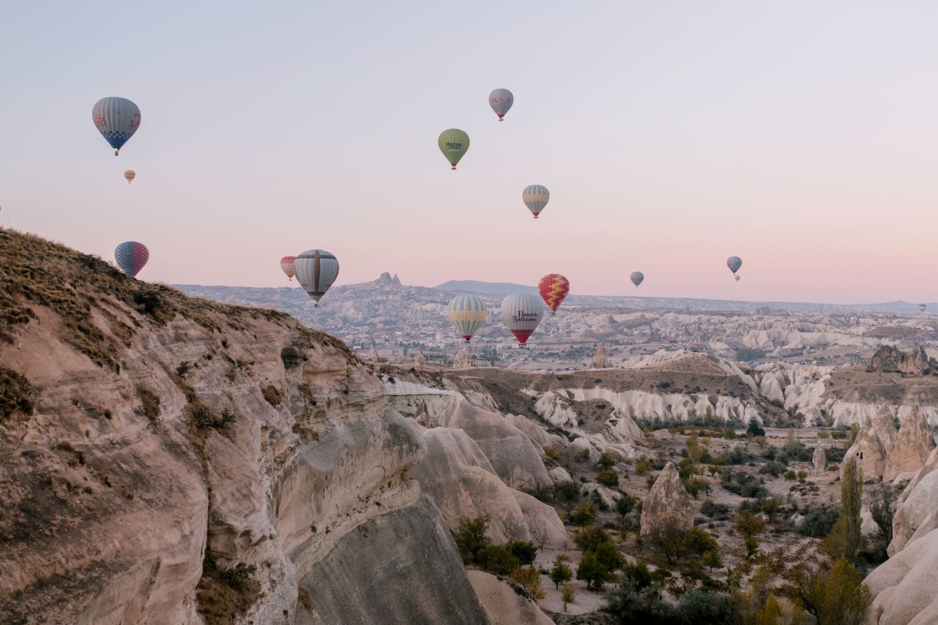 Dozens of colorful hot air balloons drifting above Cappadocia's cone-shaped fairy chimneys at sunrise, the valley glowing orange and gold below