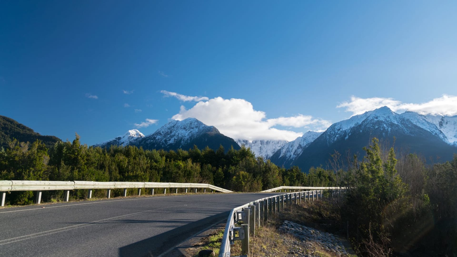 Gravel road cutting through dense Patagonian forest toward snow-capped mountains under dramatic clouds