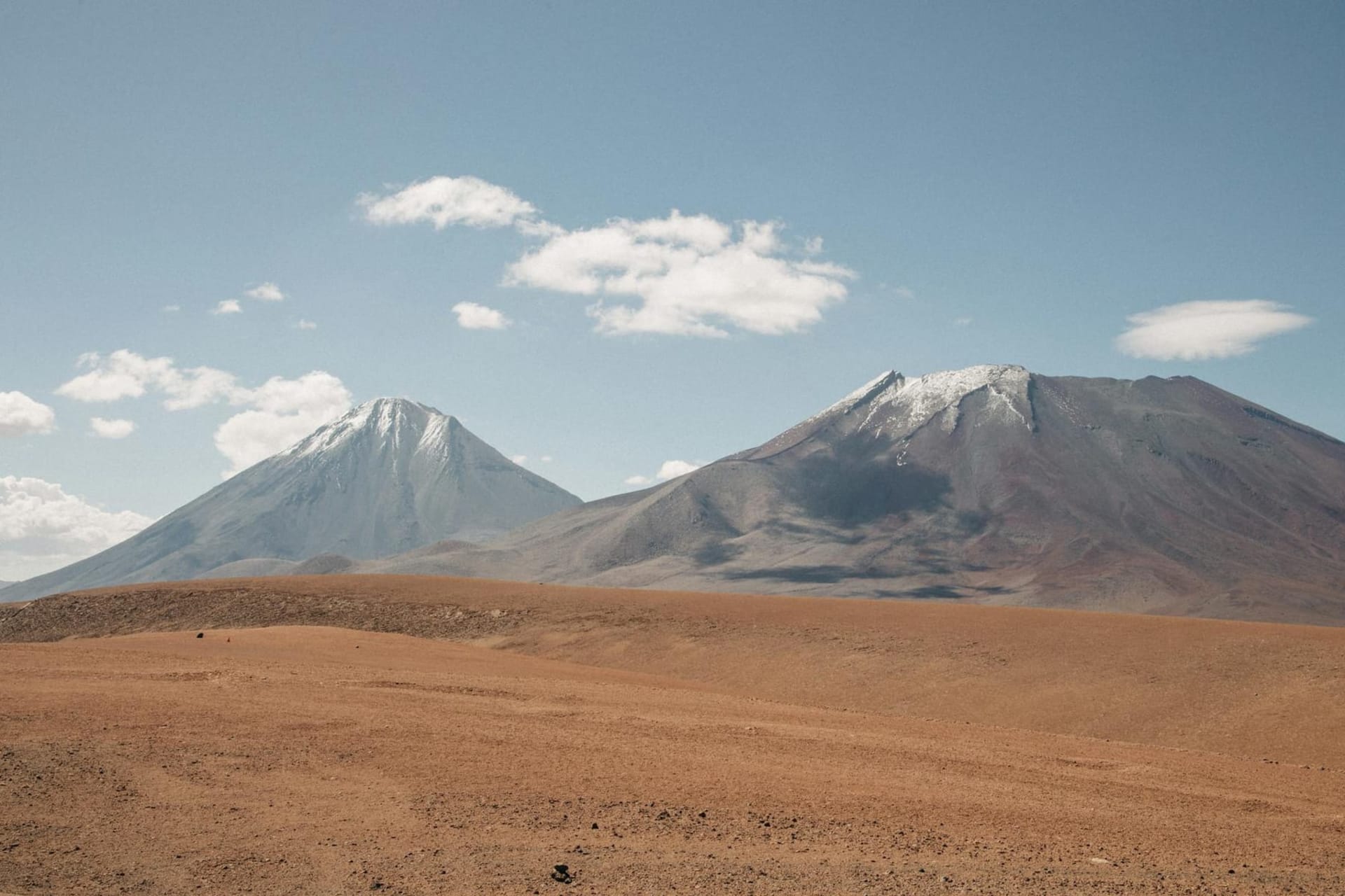 Vast white salt flats stretching to the horizon beneath a sky bruised violet and orange at dusk, with snow-capped volcanoes rising sharply in the distance across the Atacama Desert in northern Chile