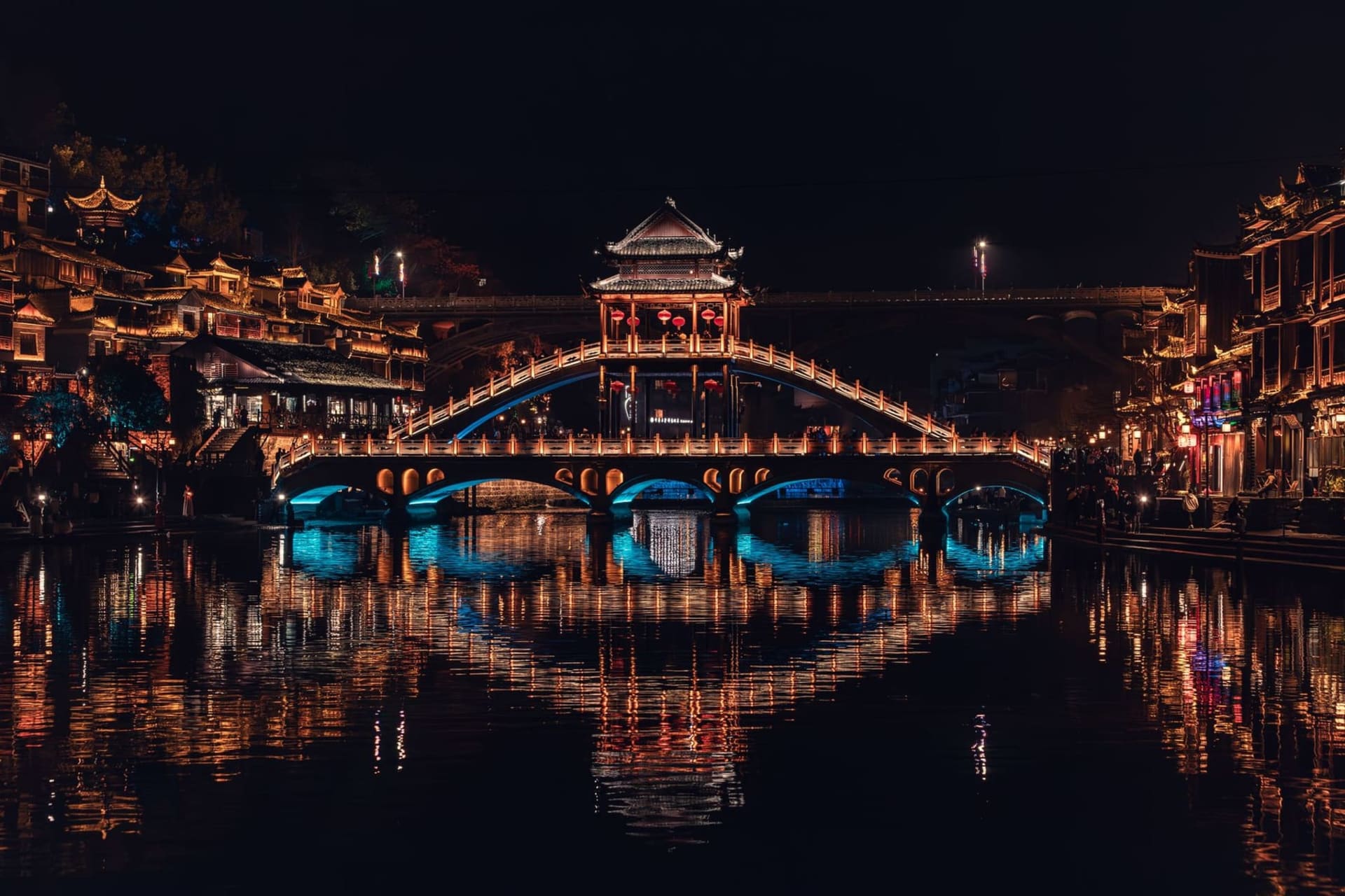 Red paper lanterns strung between stilted wooden houses overhanging the jade-green Tuojiang River at dusk, their reflections rippling in the current in Fenghuang ancient town
