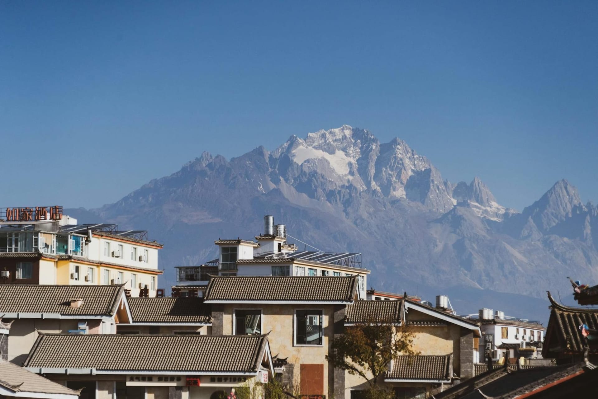 Narrow cobblestone alley in Lijiang Old Town lined with wooden Naxi architecture, red lanterns strung overhead, and a clear canal running alongside stone-paved streets with Jade Dragon Snow Mountain visible in the distant background.
