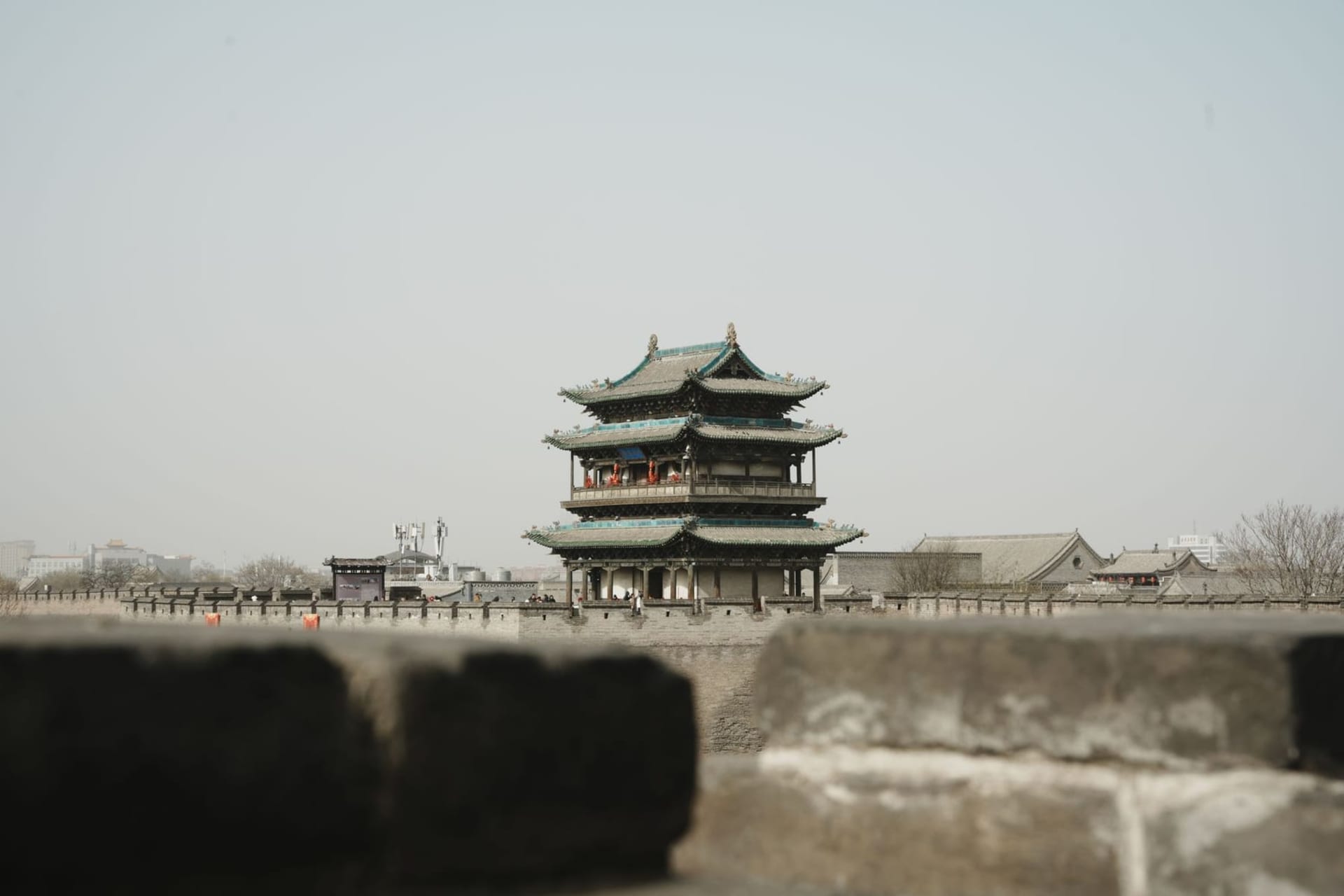 Ancient Pingyao city walls stretching across a misty dusk skyline, with Ming-dynasty watchtowers and grey-tiled rooftops below illuminated by warm lantern light