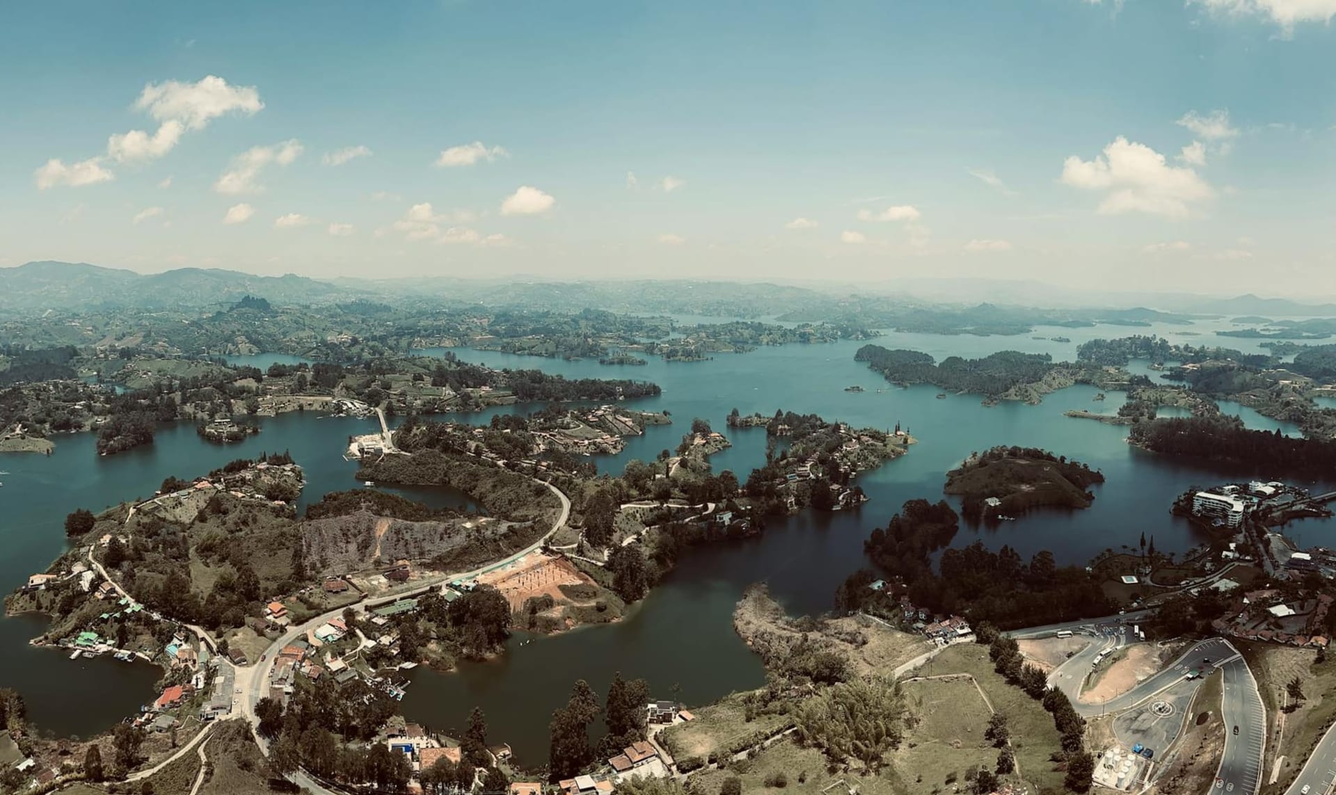 The Piedra del Peñol monolith rising dramatically above the blue-green reservoir at Guatapé, its sheer granite face glowing amber in afternoon light with a white zigzag staircase visible cutting up through a narrow crack in the rock.
