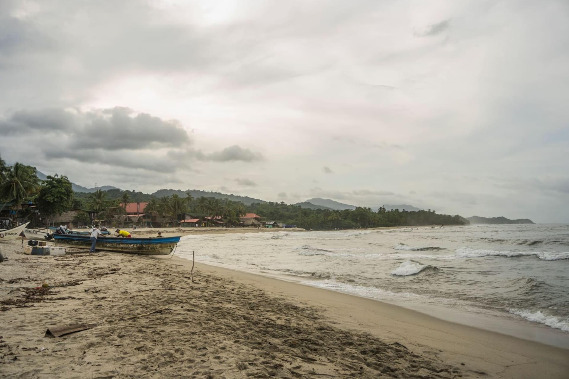 A wide, brown jungle river flowing between dense tropical vegetation toward a white-sand beach where it opens into the turquoise Caribbean Sea, with the hazy peaks of the Sierra Nevada rising in the distance.