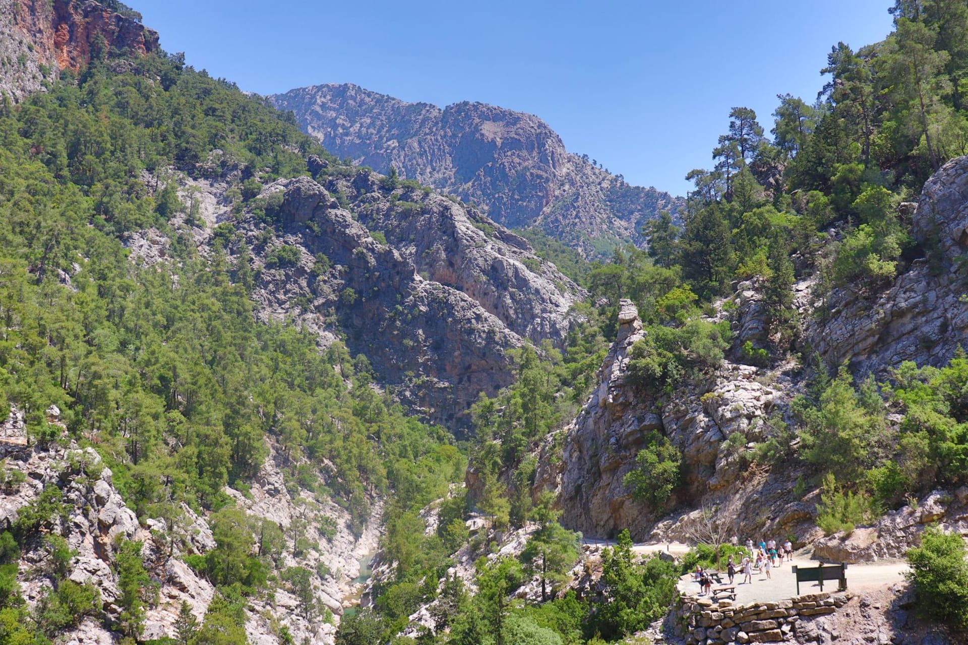Hikers on a rocky mountain trail through a lush Cretan gorge under clear blue skies