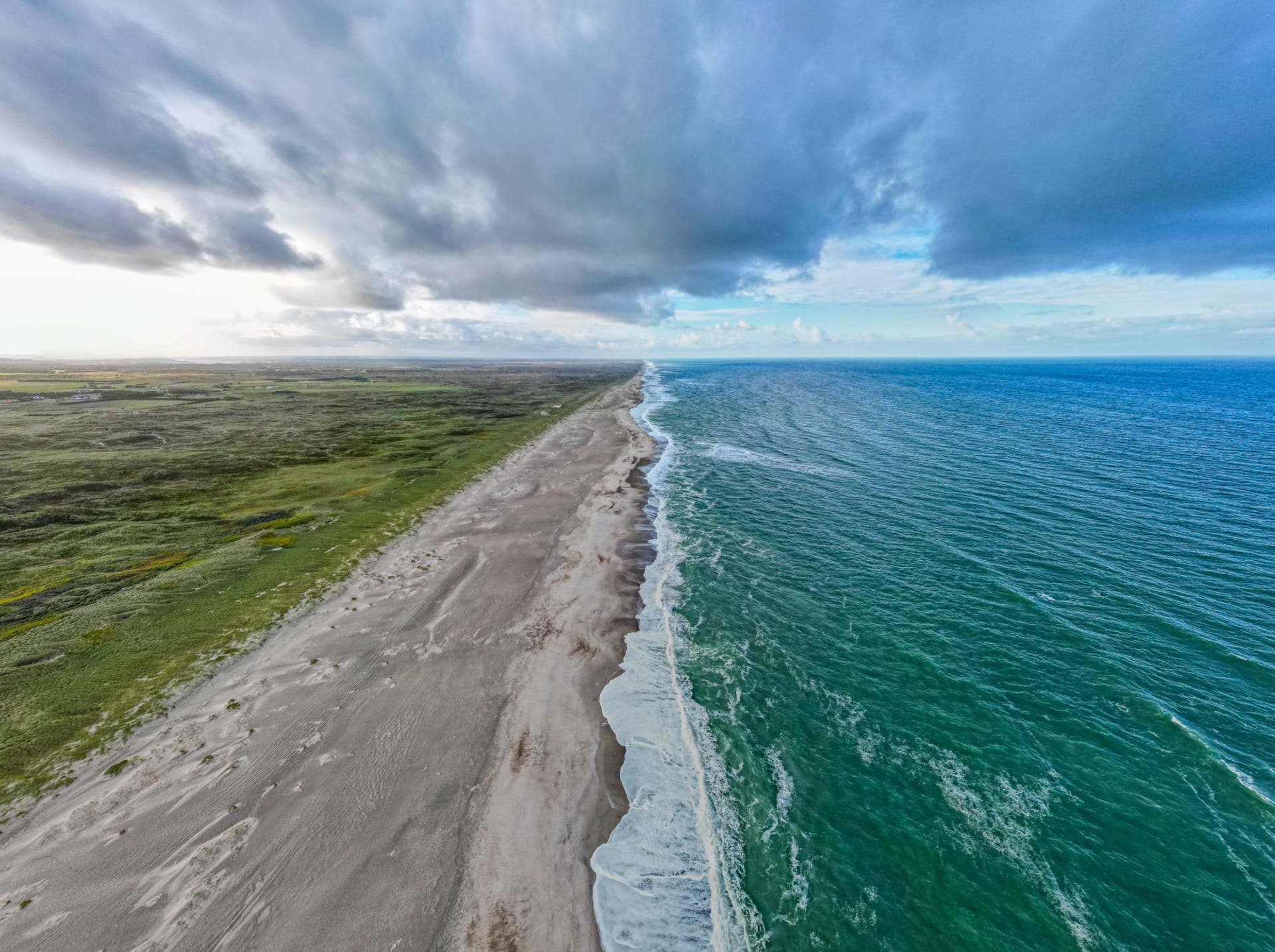 Aerial view of the dramatic Danish coastline in Thisted, with pale sand beach, deep blue sea, and sweeping storm clouds overhead