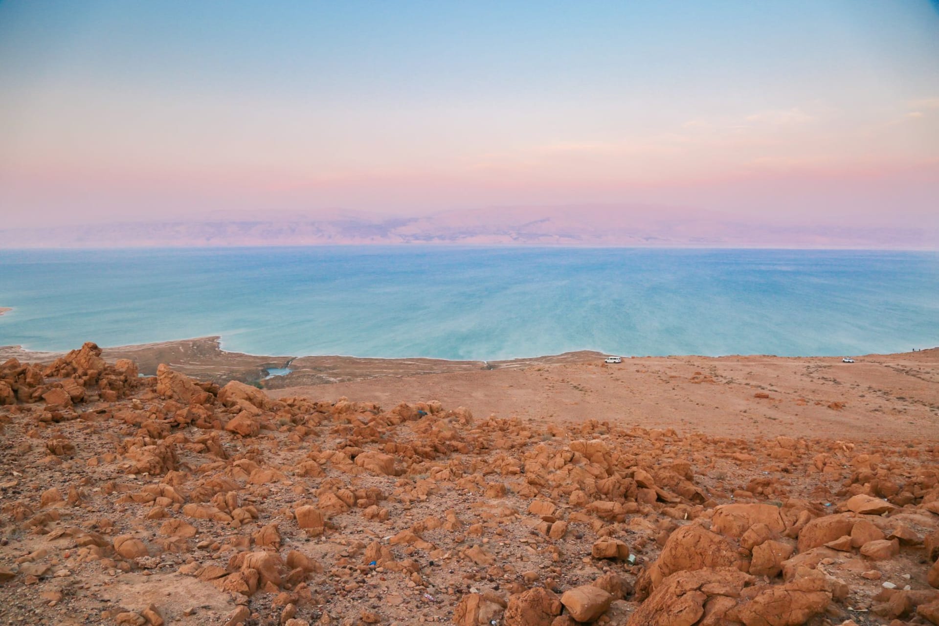 Panoramic view of the Dead Sea at sunset, rocky shoreline in the foreground and a pastel pink and orange sky stretching over the still, hazy water