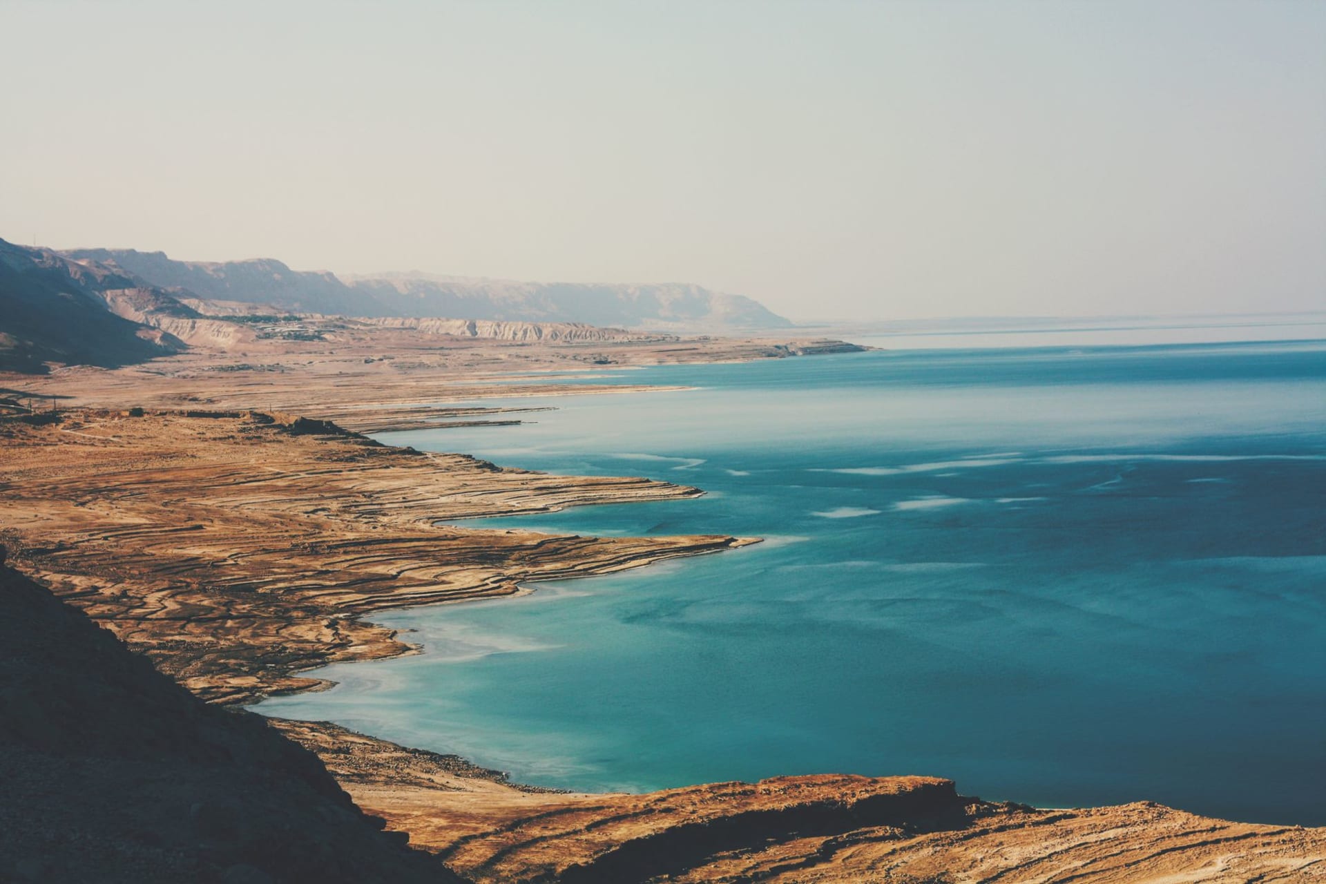 The pale limestone cliffs of Qumran with the dark openings of caves visible above the ancient settlement ruins, the Dead Sea glinting in the distance