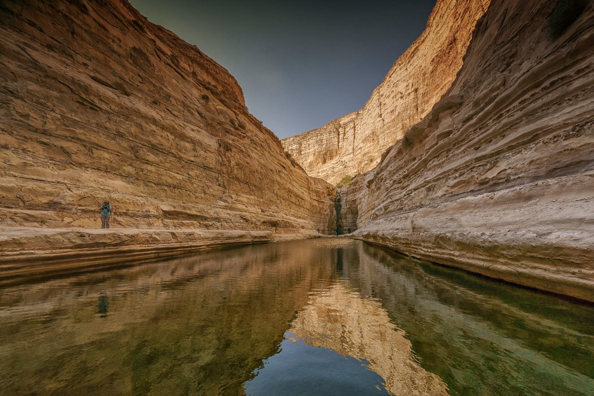 A narrow red-rock slot canyon in Wadi Mujib with turquoise river water rushing between towering cliff walls, shafts of light from above