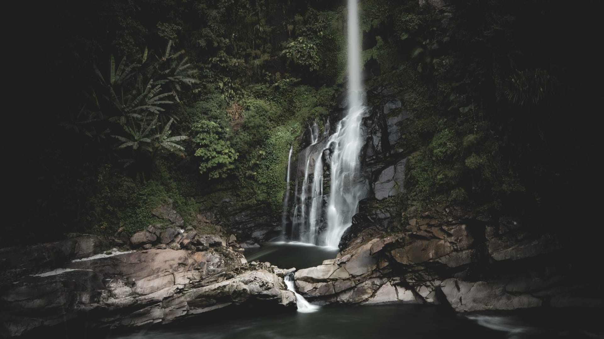 Two waterfalls cascading side by side over dark volcanic rock into a pool, surrounded by dense green rainforest