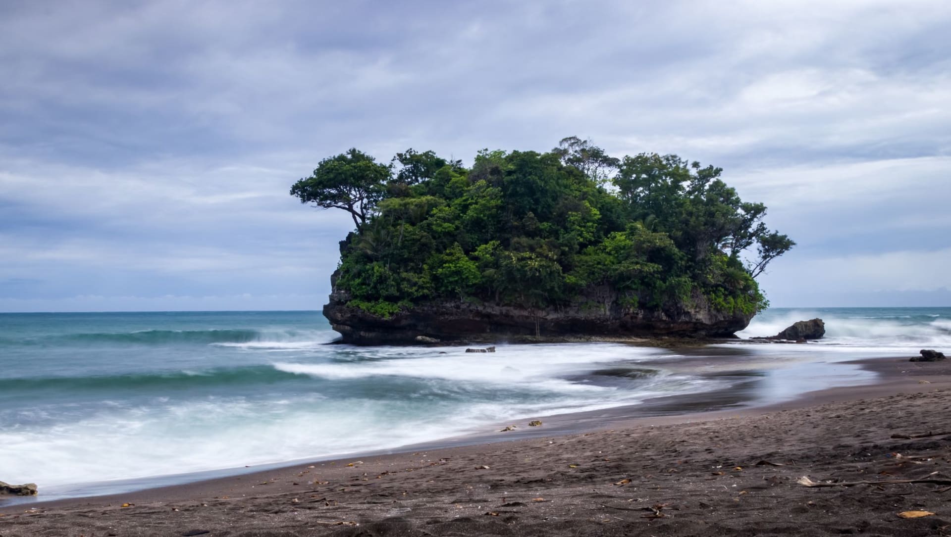 Lush tropical island rising from calm ocean waters under a hazy sky, dense greenery covering the slopes
