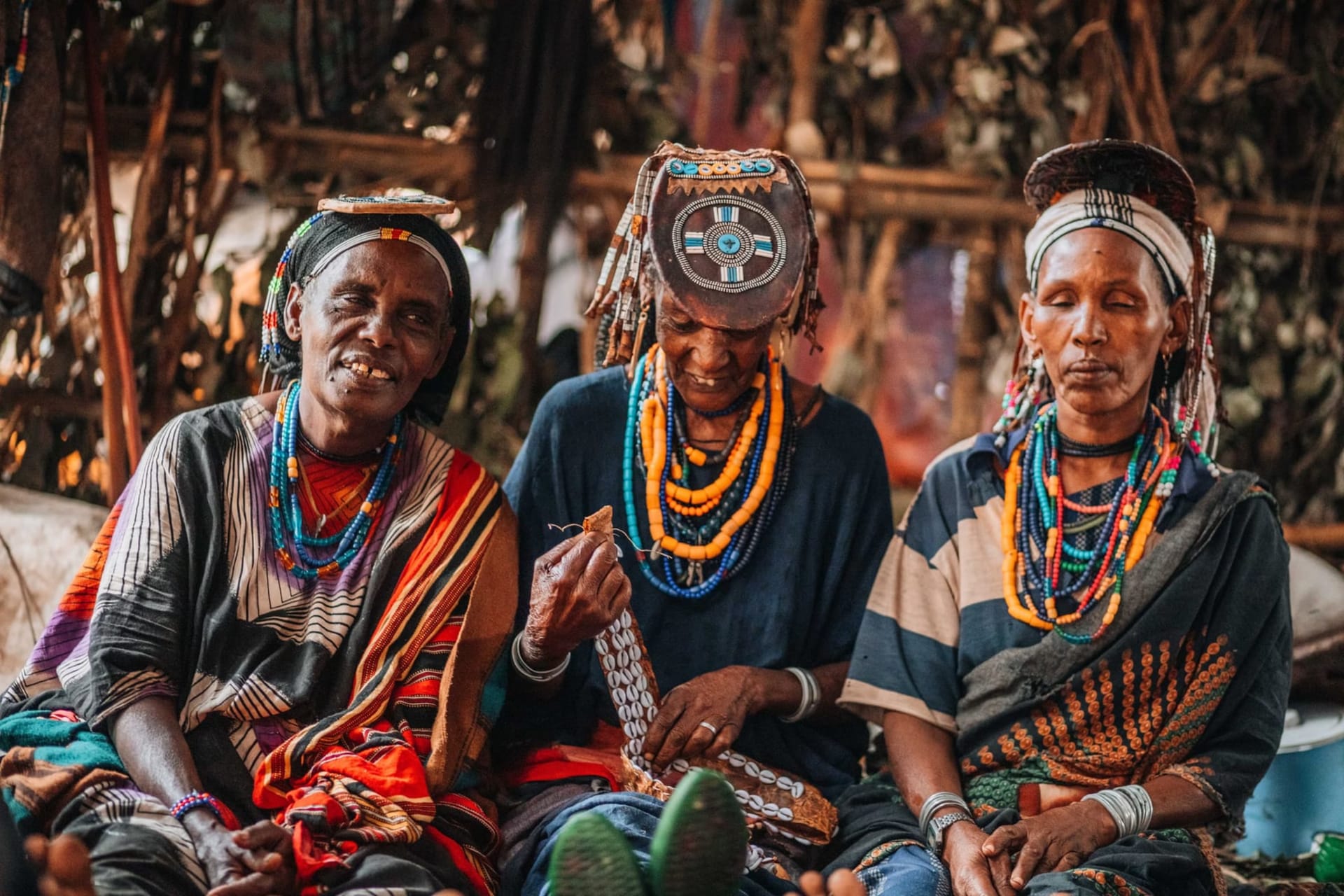 A Hamar woman with ochre-painted skin and metal neck rings stands before a dry acacia landscape, her beaded chest ornaments catching the low afternoon light of the Omo Valley.