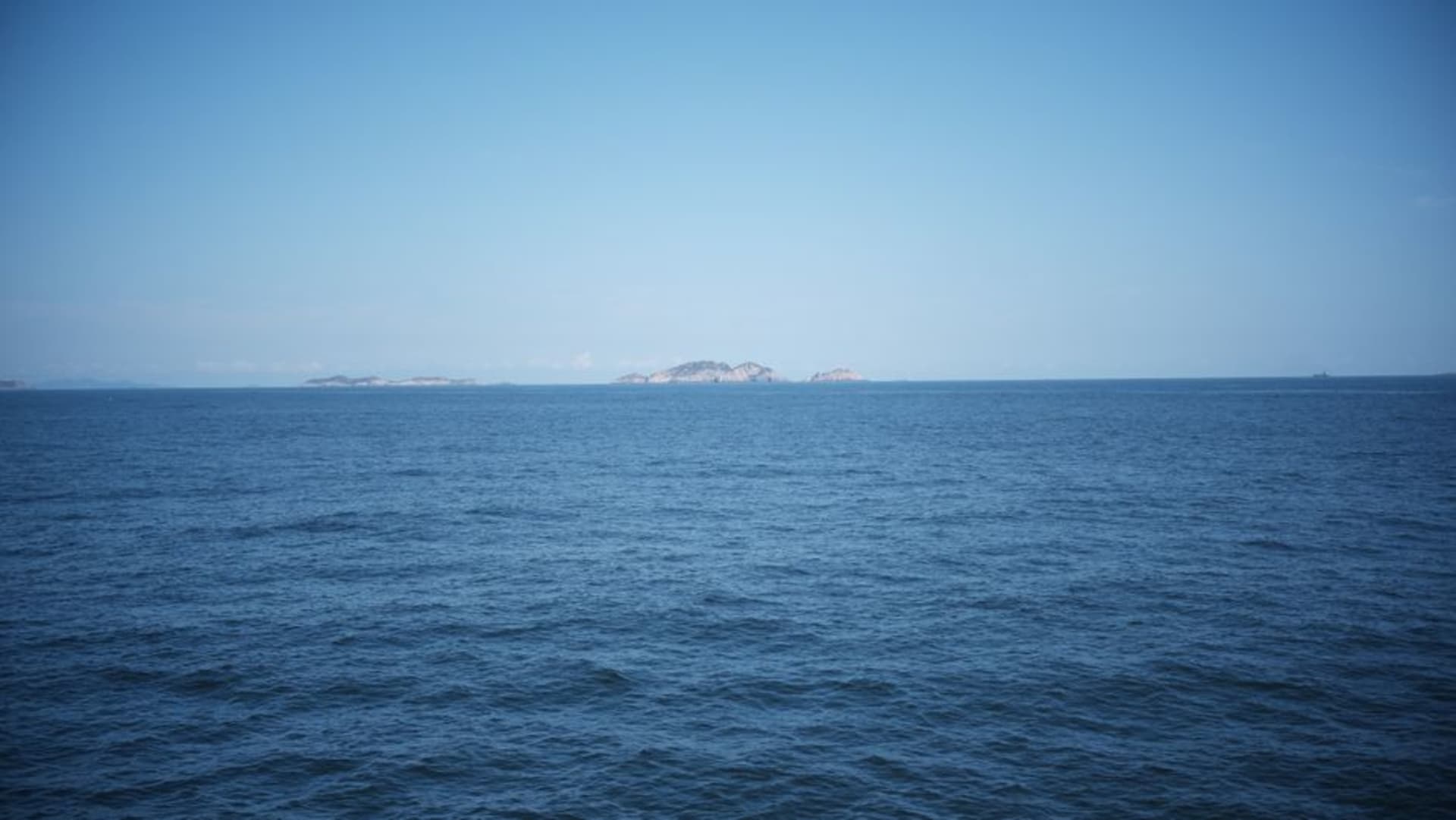 A forested hillside island rising from a calm turquoise bay, with a narrow strip of white sand beach at its base