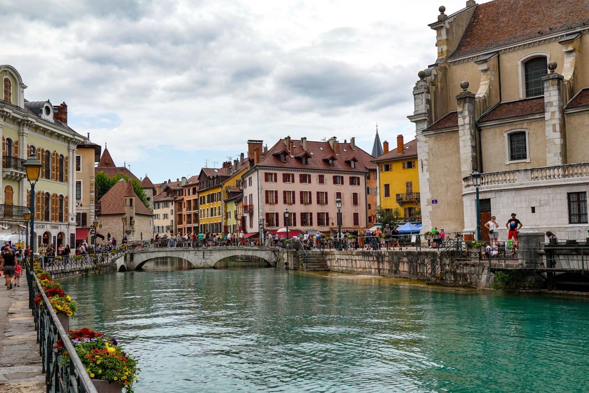 The turquoise water of Lac d'Annecy with the old town's pastel buildings along a canal and snow-streaked Alps rising behind