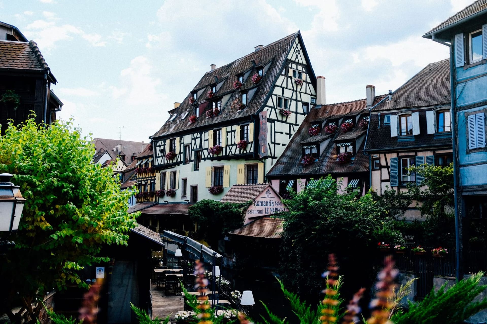 Pastel half-timbered houses in coral, sage, and ochre lining a narrow canal in Colmar's Petite Venise quarter, their reflections rippling in the still green water beneath flower-draped balconies