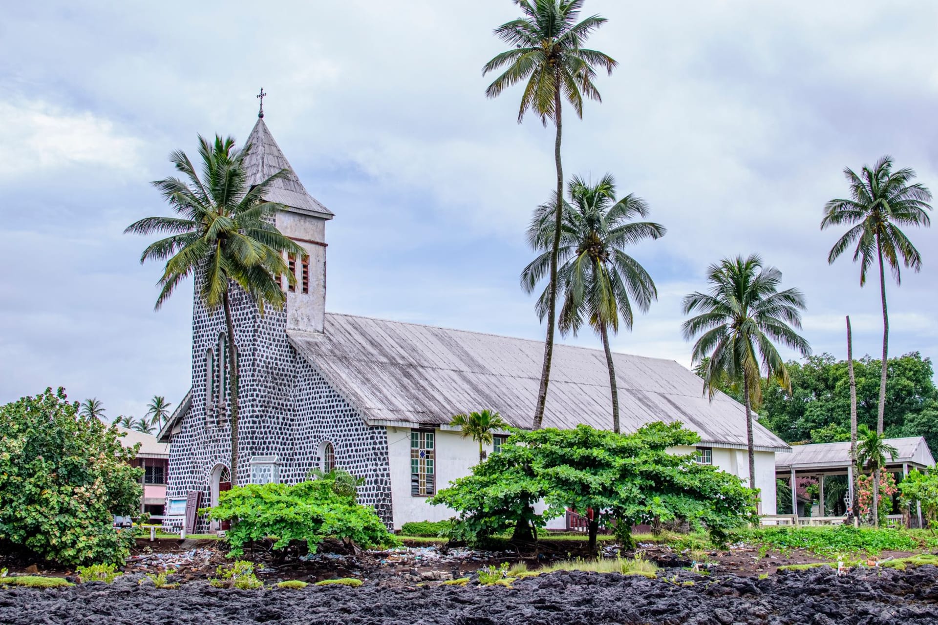 The weathered colonial facade of the Camp de la Transportation in Saint-Laurent-du-Maroni at golden hour, with a palm tree casting shadow across the old prison walls
