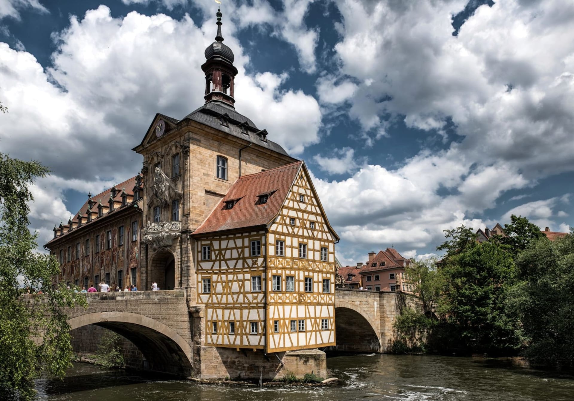The Bamberg Old Town Hall perched on an artificial island in the Regnitz river, its painted baroque facade reflected in the dark water, with the spires of the cathedral rising behind it on the hill