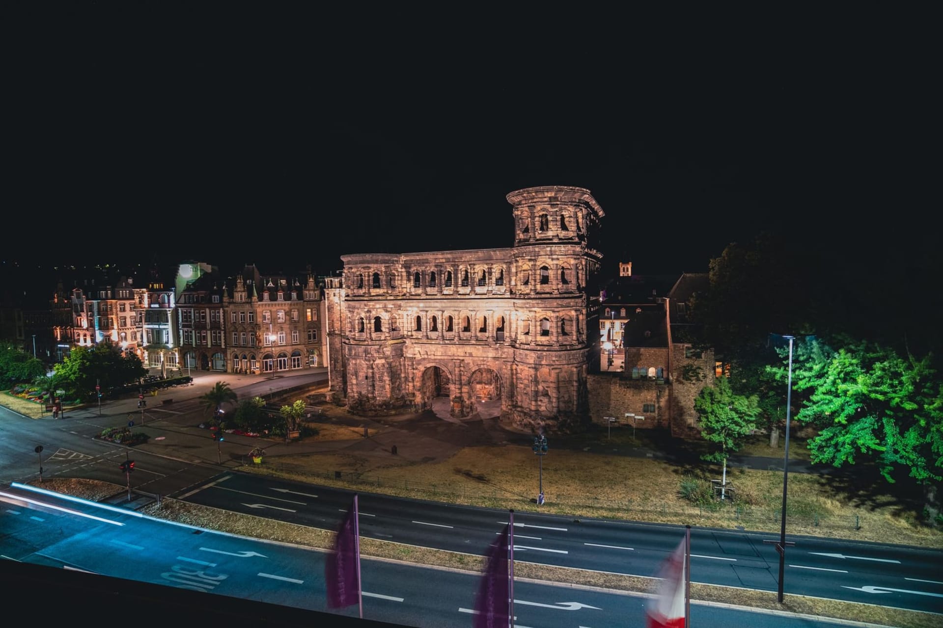 The Porta Nigra, Trier's massive blackened Roman gate, rising above a cobblestone square on a grey-blue afternoon, pedestrians dwarfed beneath its ancient arches